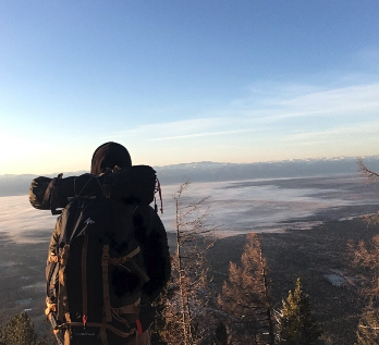Un randonneur avec un sac à dos observe un paysage de montagne et de forêt sous un ciel clair.