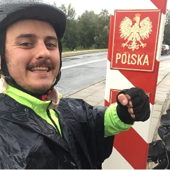 Jeune homme souriant en vêtements de pluie, devant un panneau indiquant la frontière polonaise avec un aigle blanc sur fond rouge et le mot 'POLSKA'.