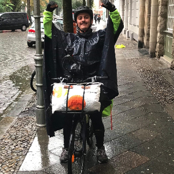 Un homme souriant à vélo sous la pluie, portant un imperméable noir et recevant une livraison de nourriture dans une ville