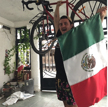 Une personne souriante tenant un drapeau mexicain, avec un vélo suspendu au plafond, dans un espace intérieur.