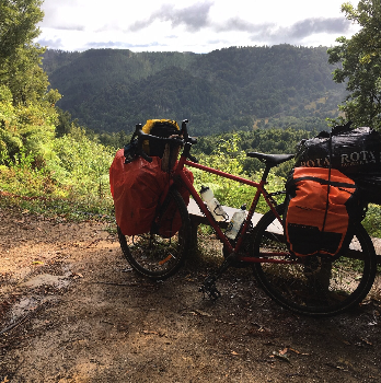 Une bicyclette rouge équipée de sacoches orange, stationnée sur un chemin de terre avec un paysage de montagnes et de forêt en arrière-plan.