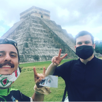 Deux hommes prennent un selfie devant une grande pyramide maya en pierre à Yucatan, l'un avec un masque noir et l'autre portant des lunettes de soleil.
