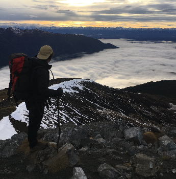 Une personne avec un sac à dos et un bâton de marche regarde le paysage de montagnes et de nuages au lever ou coucher du soleil, avec de la neige et des rochers autour.