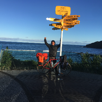Une personne avec un vélo rouge devant un panneau de direction coloré près de la mer, levant un bras en signe de triomphe, avec un ciel clair et un paysage côtier en arrière-plan.