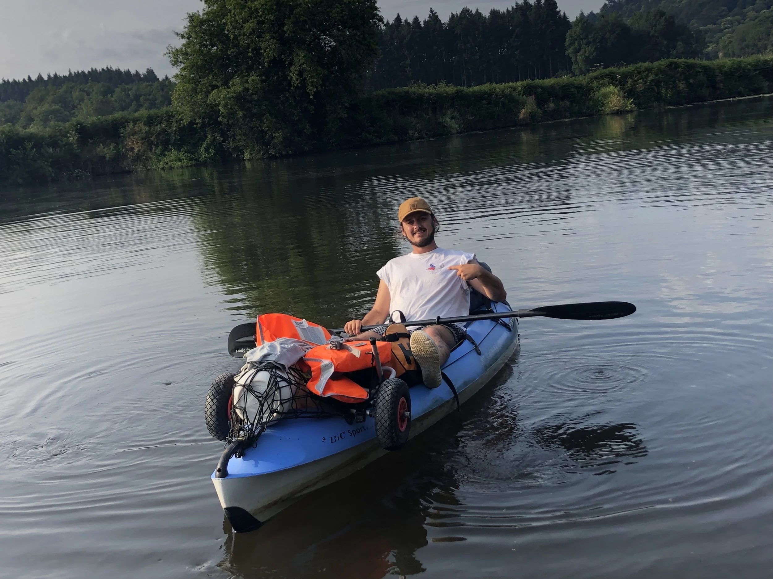 Une personne en kayak sur une rivière entourée d'arbres et de nature, souriant et pointant du doigt quelque chose.