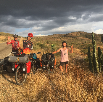 Deux personnes à vélo avec des sacs de voyage, une femme debout à côté d'eux faisant un signe de victoire, dans un paysage désertique avec des cactus et un ciel nuageux.