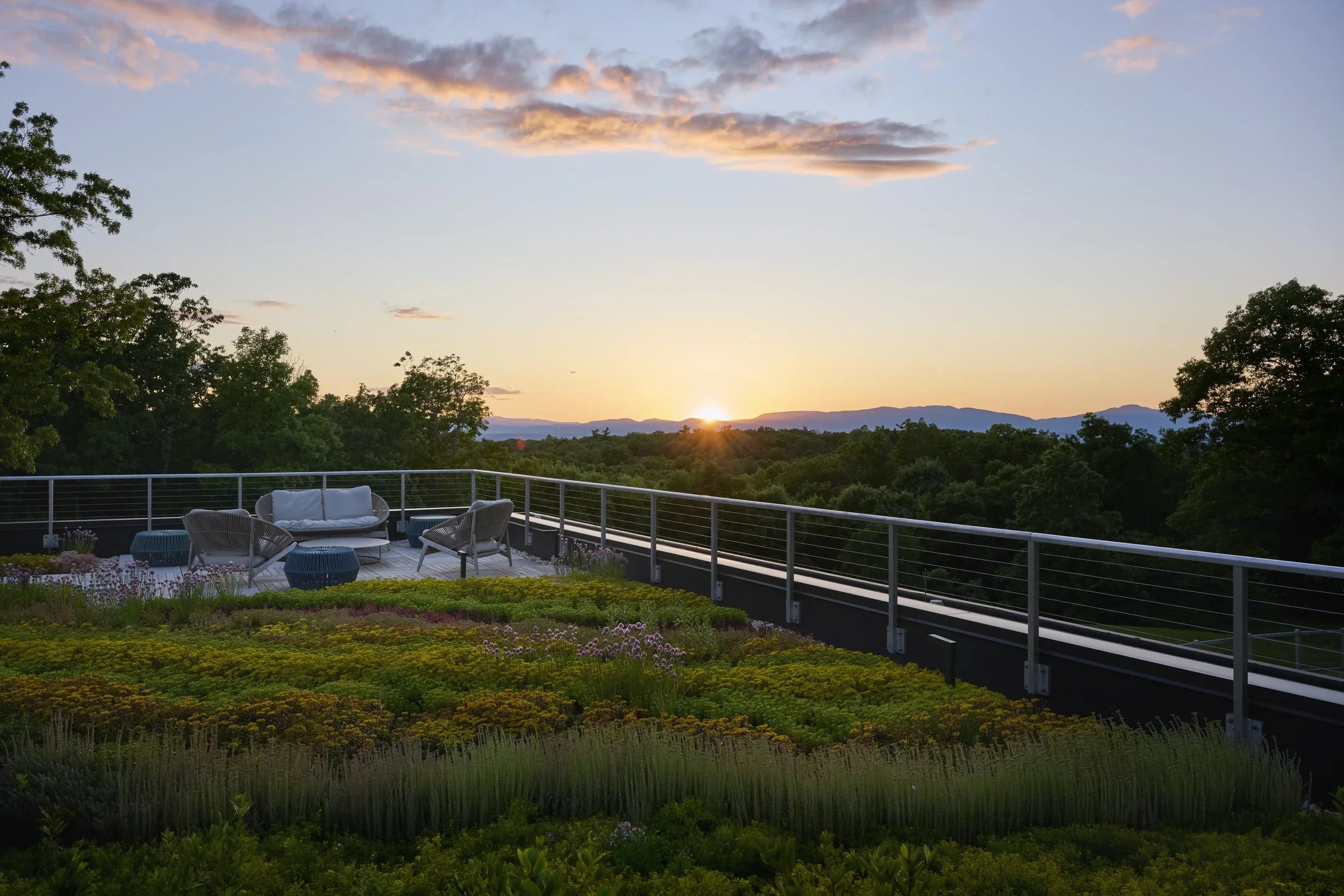 Sunset view from a rooftop patio with outdoor furniture, overlooking a green landscape with trees and distant mountains.