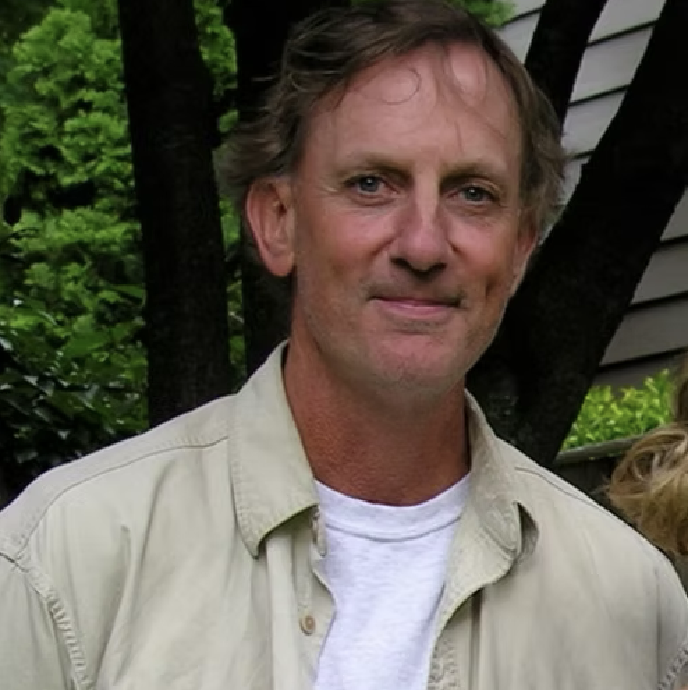 A middle-aged man with light brown hair and blue eyes, wearing a beige button-up shirt over a white t-shirt, standing outdoors near a tree and house with green foliage in the background.