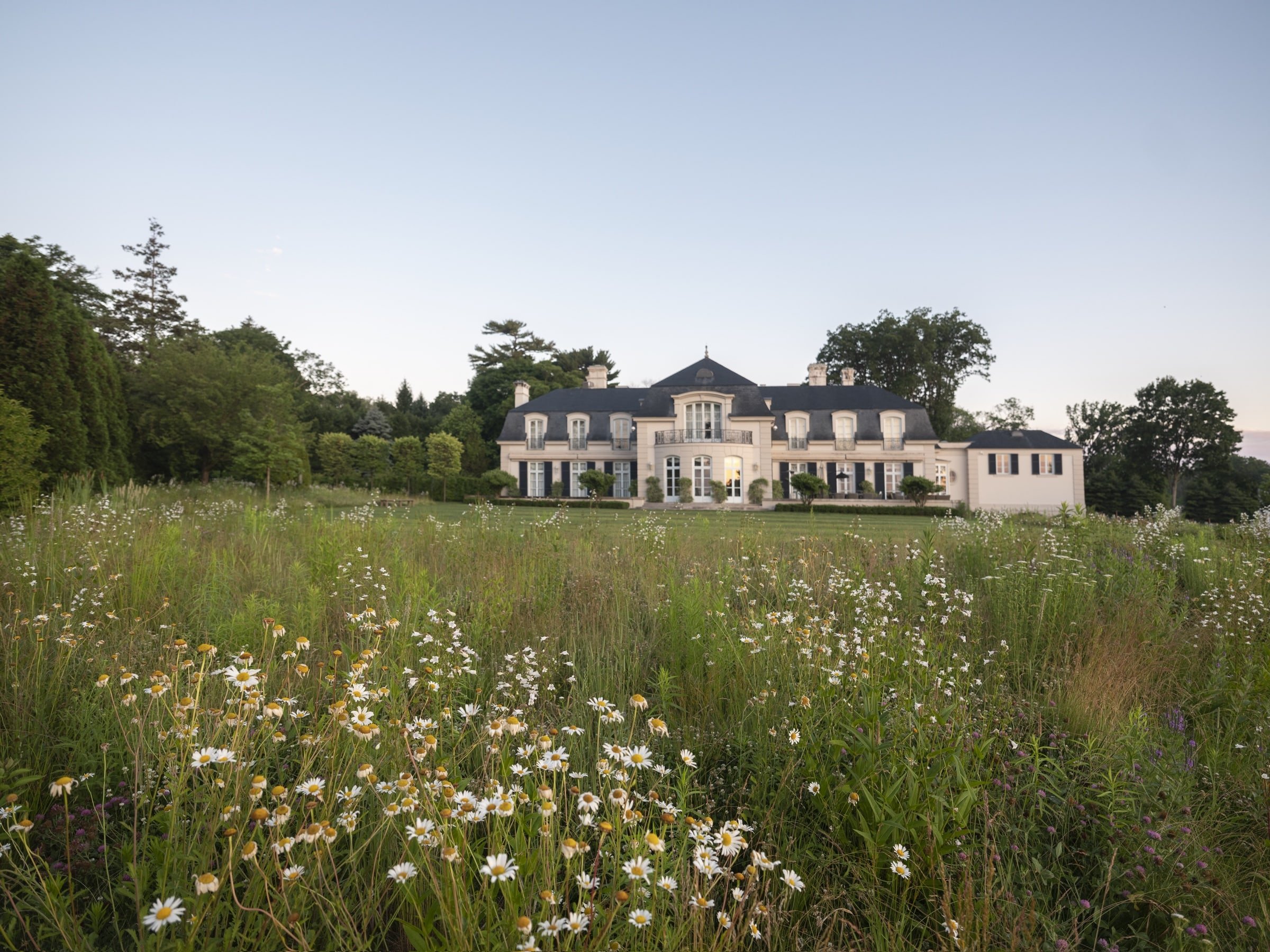 A large elegant white mansion with dark roofing, surrounded by a lush garden and tall trees, under a clear sky.