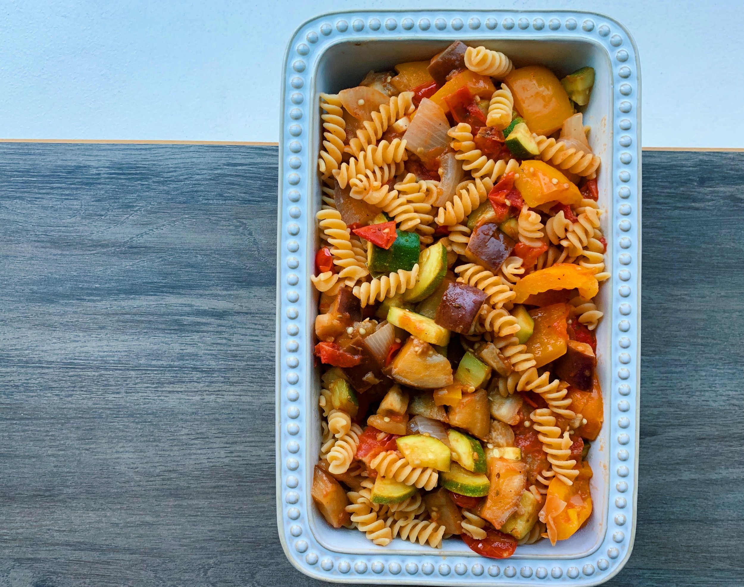 White rectangular dish filled with rotini pasta mixed with assorted vegetables like zucchini, bell peppers, onions, and tomatoes.