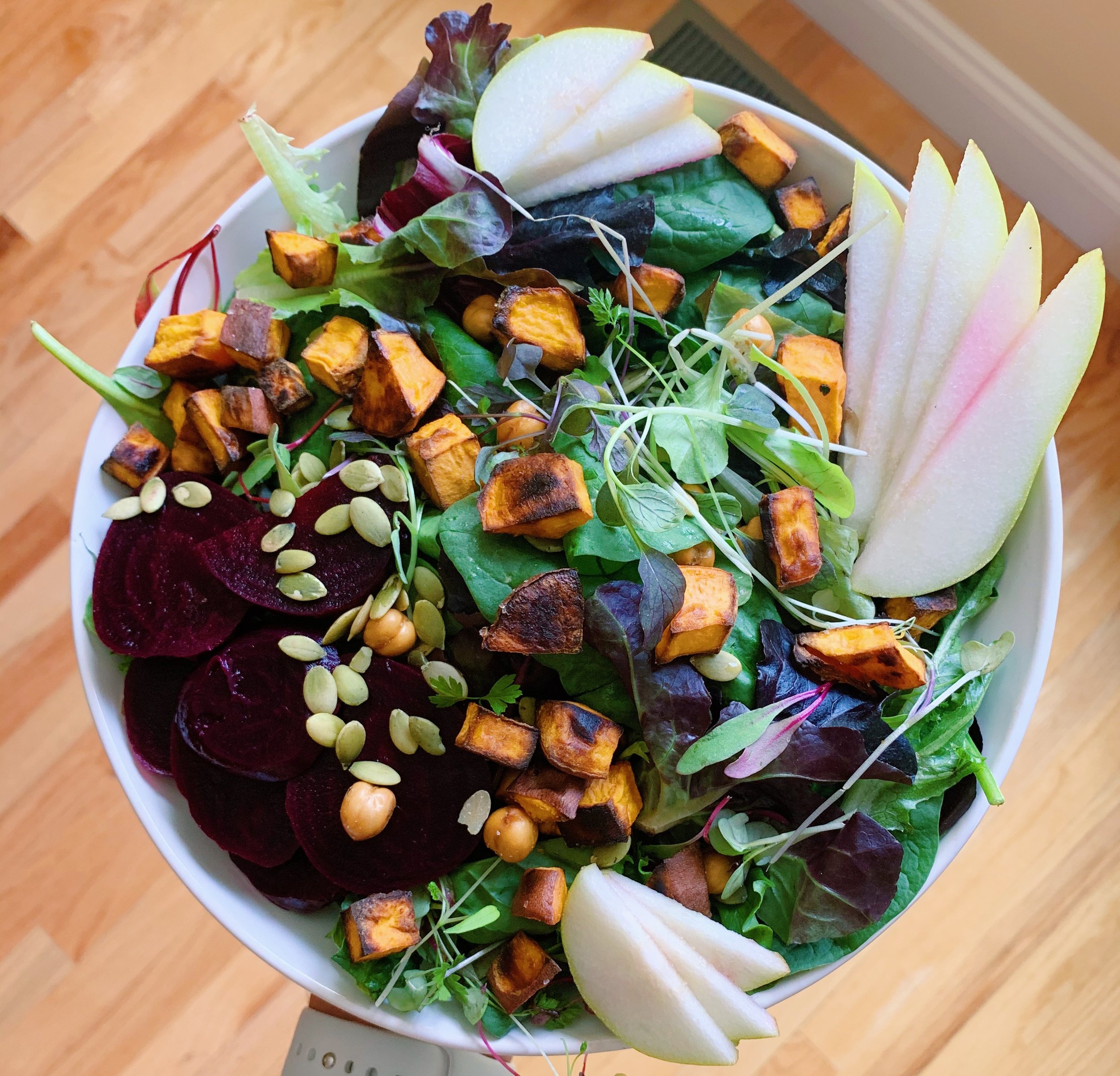 A salad with mixed greens, sliced apples, roasted sweet potatoes, beets, chickpeas, pumpkin seeds, and microgreens in a white bowl on a wooden surface.