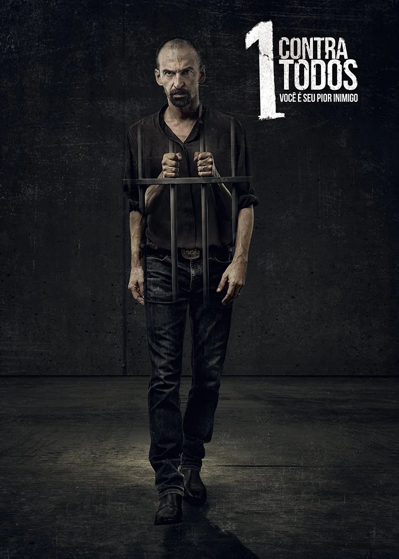 Man standing behind prison bars in dark room with text "1 Contra Todos: Você é seu pior inimigo" on wall.