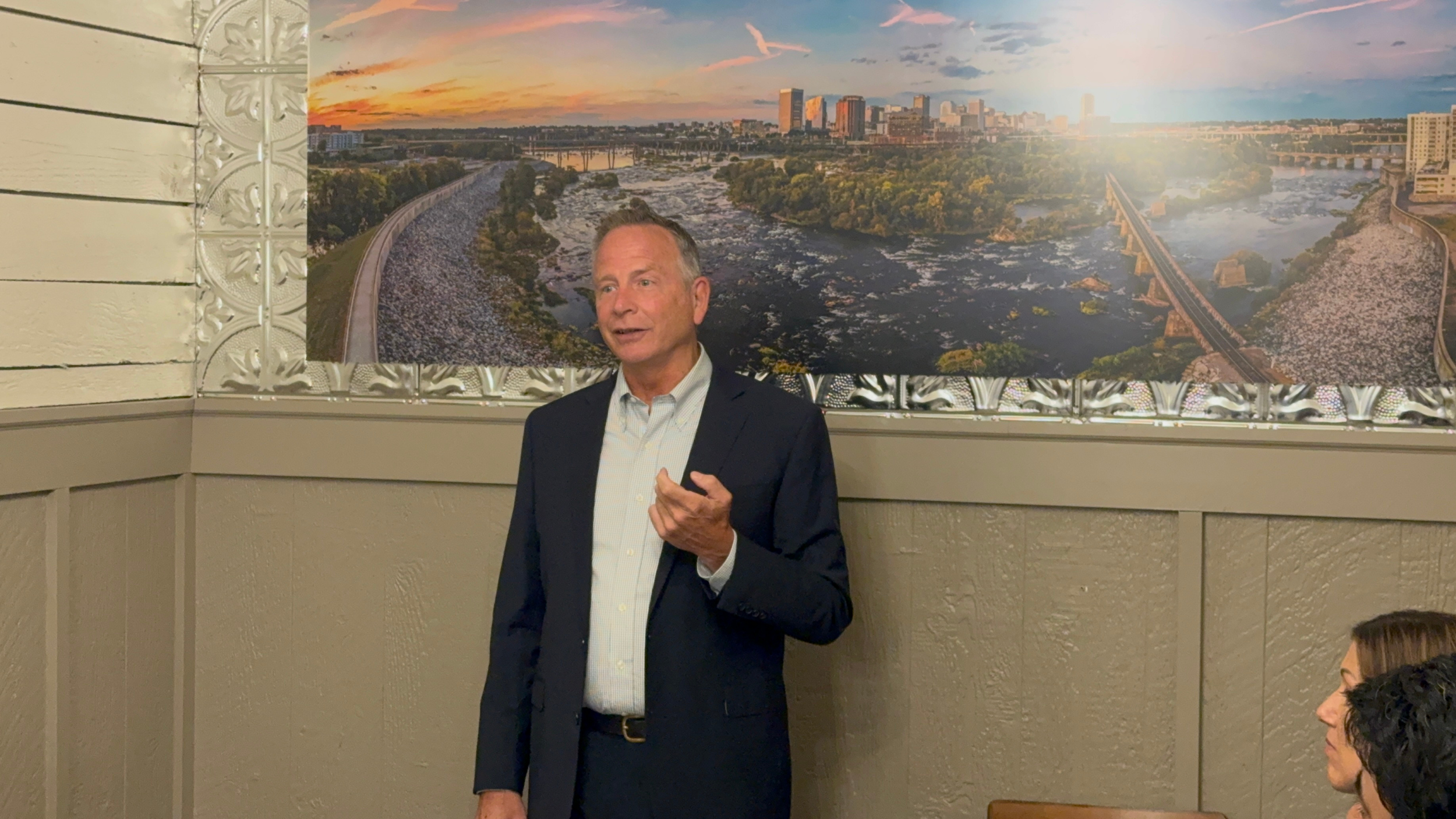 A man in a dark suit and light shirt speaking in front of a large landscape mural of a city and river at sunset, with a woman listening in the foreground.