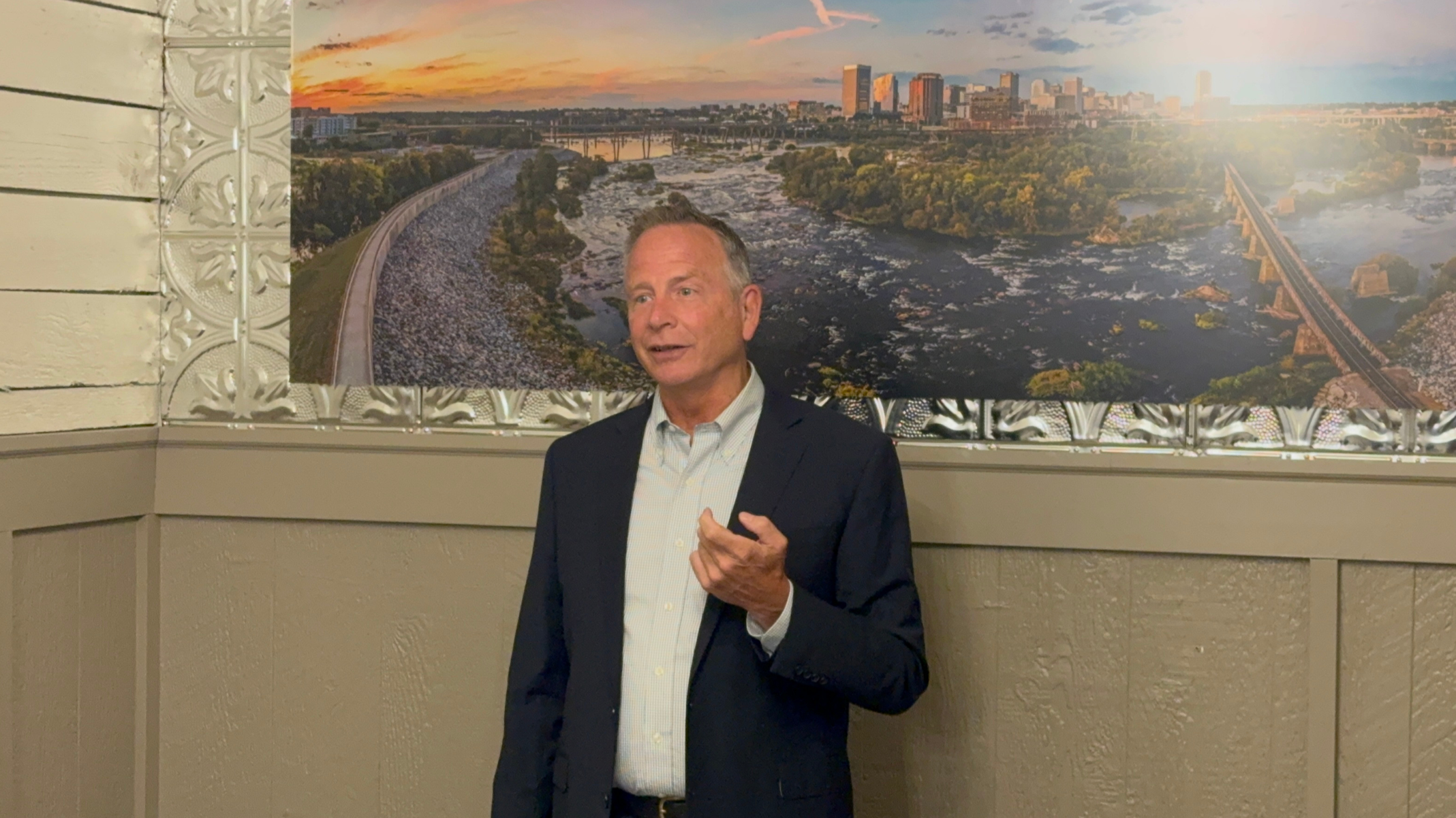 A man in a suit and light shirt standing indoors in front of a large aerial cityscape photograph with a river, bridges, and buildings during sunset.