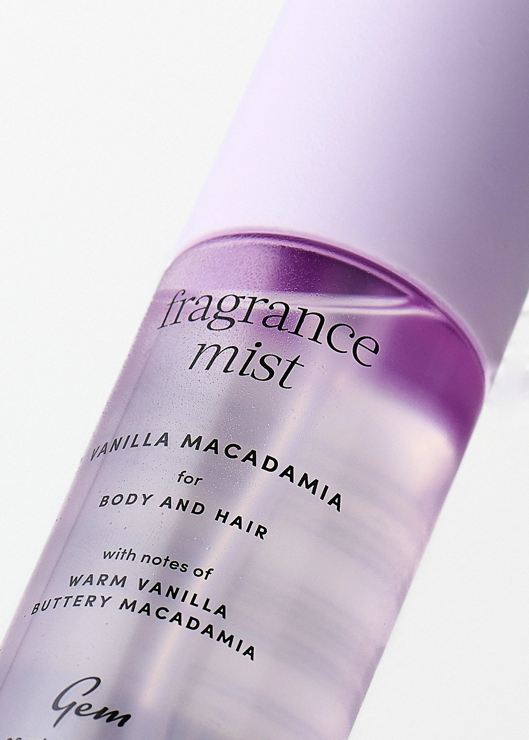 Close-up of a lavender-colored body mist spray bottle labeled 'fragrance mist' with additional text about vanilla and macadamia notes for body and hair, set against a white background.