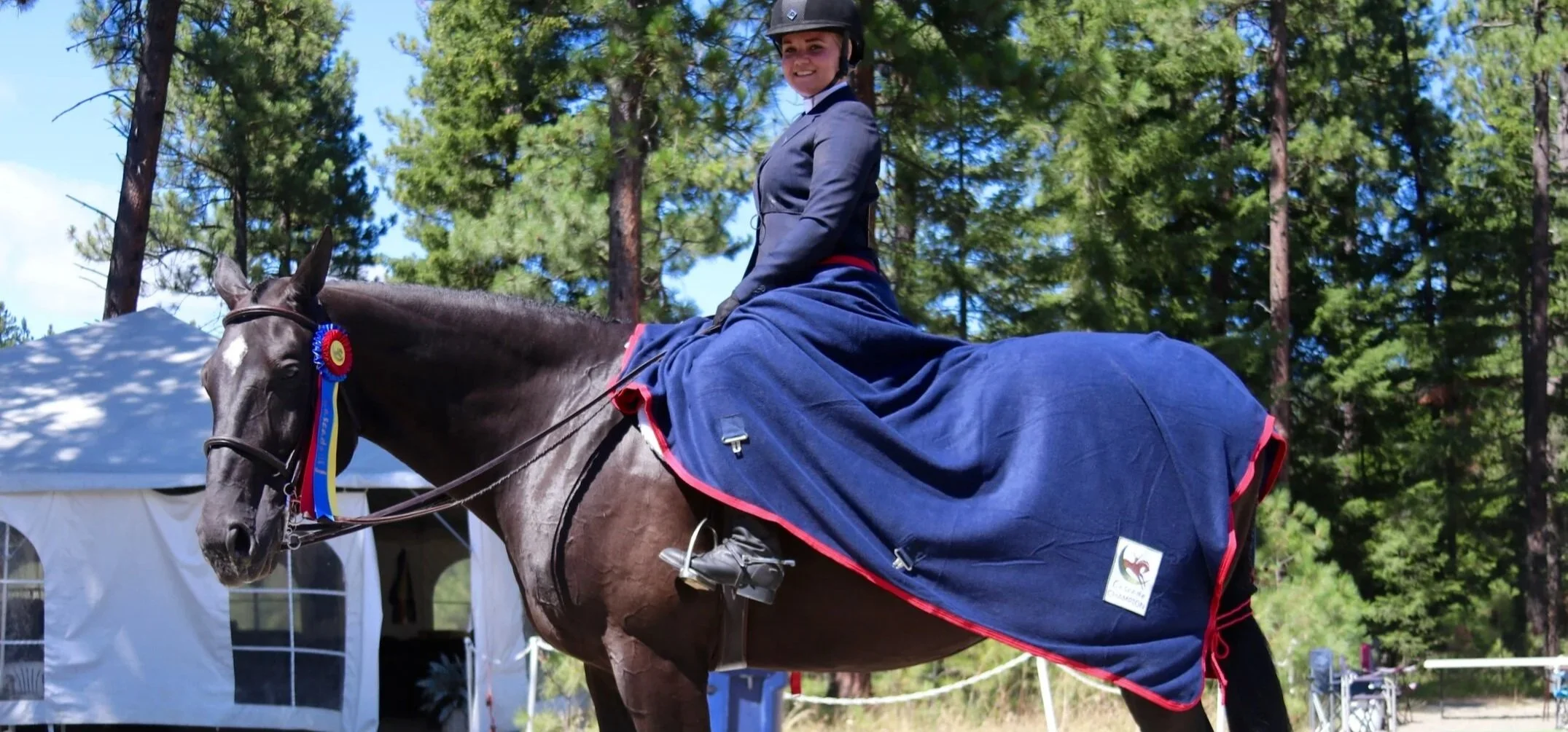 A woman in formal riding attire, including a helmet, riding a dark brown horse with a white star on its forehead at an outdoor equestrian event in a wooded area. The horse has a blue blanket with red trim and a ribbon indicating an award.