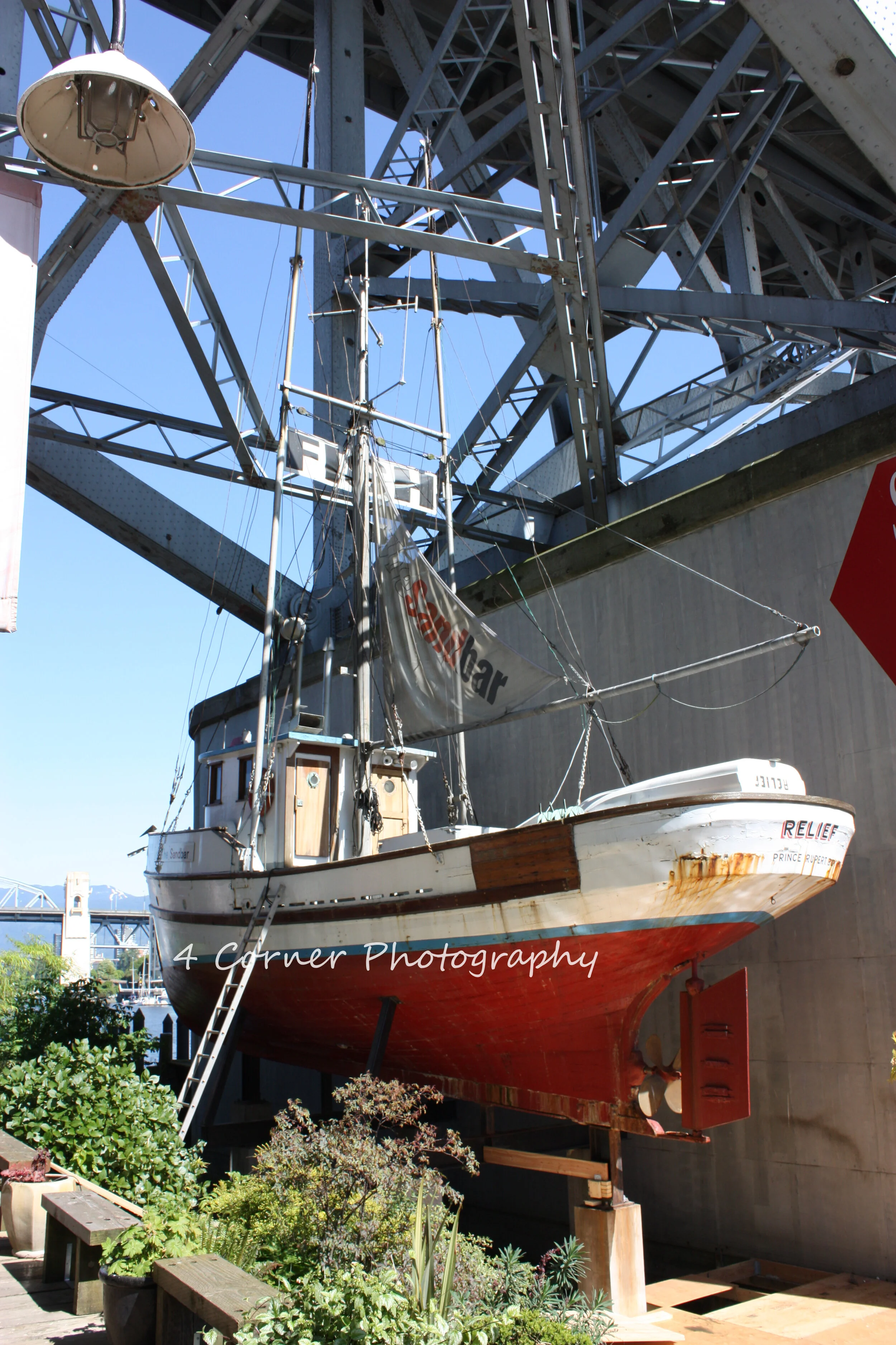 A small boat named 'RELIEF' is displayed on a stand beneath a large railway bridge, with plants at the foreground.