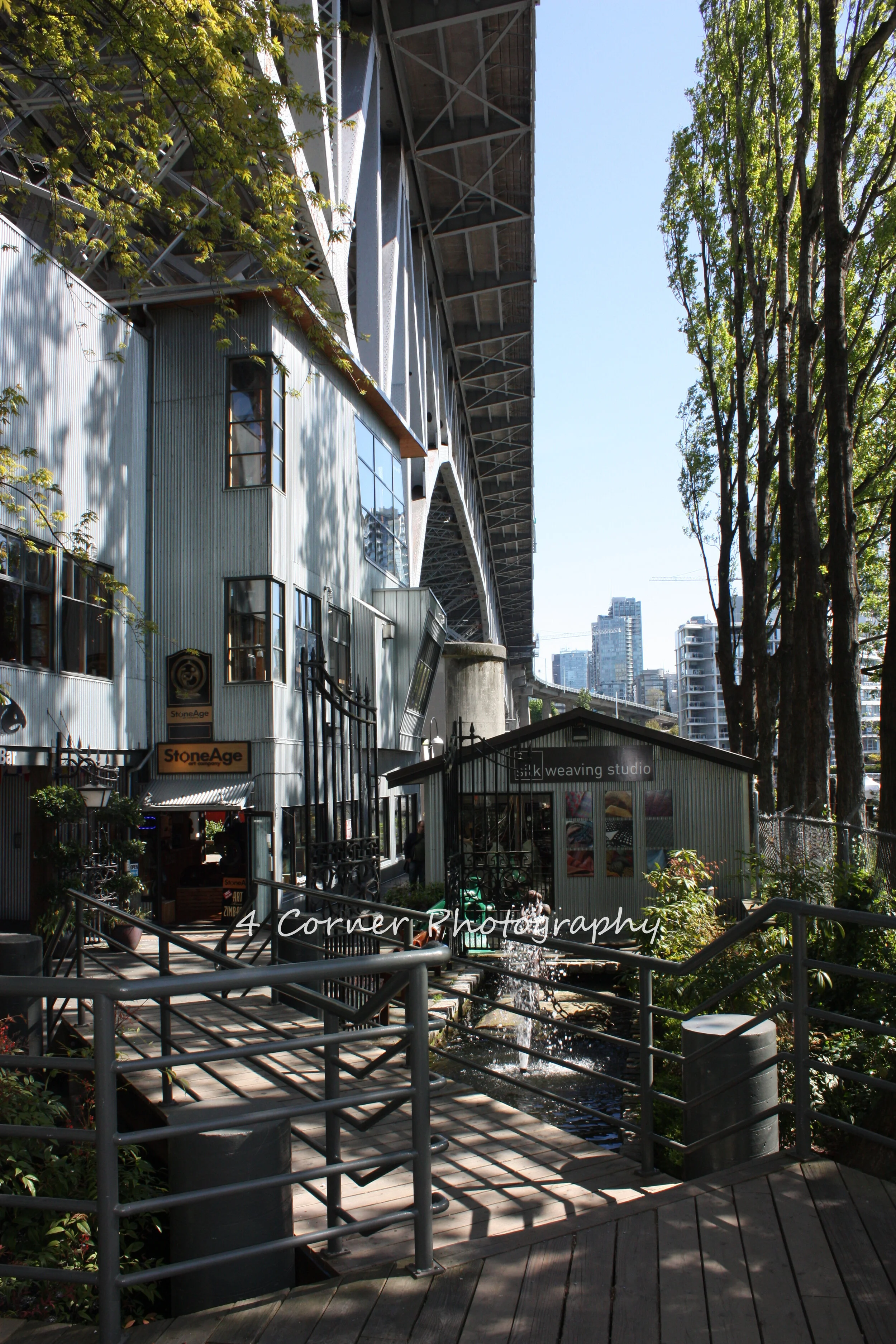 An urban scene with a small, corrugated metal building housing a weaving studio, a narrow water feature with a stepping stone, surrounded by trees and city buildings in the background, with a large bridge overhead.