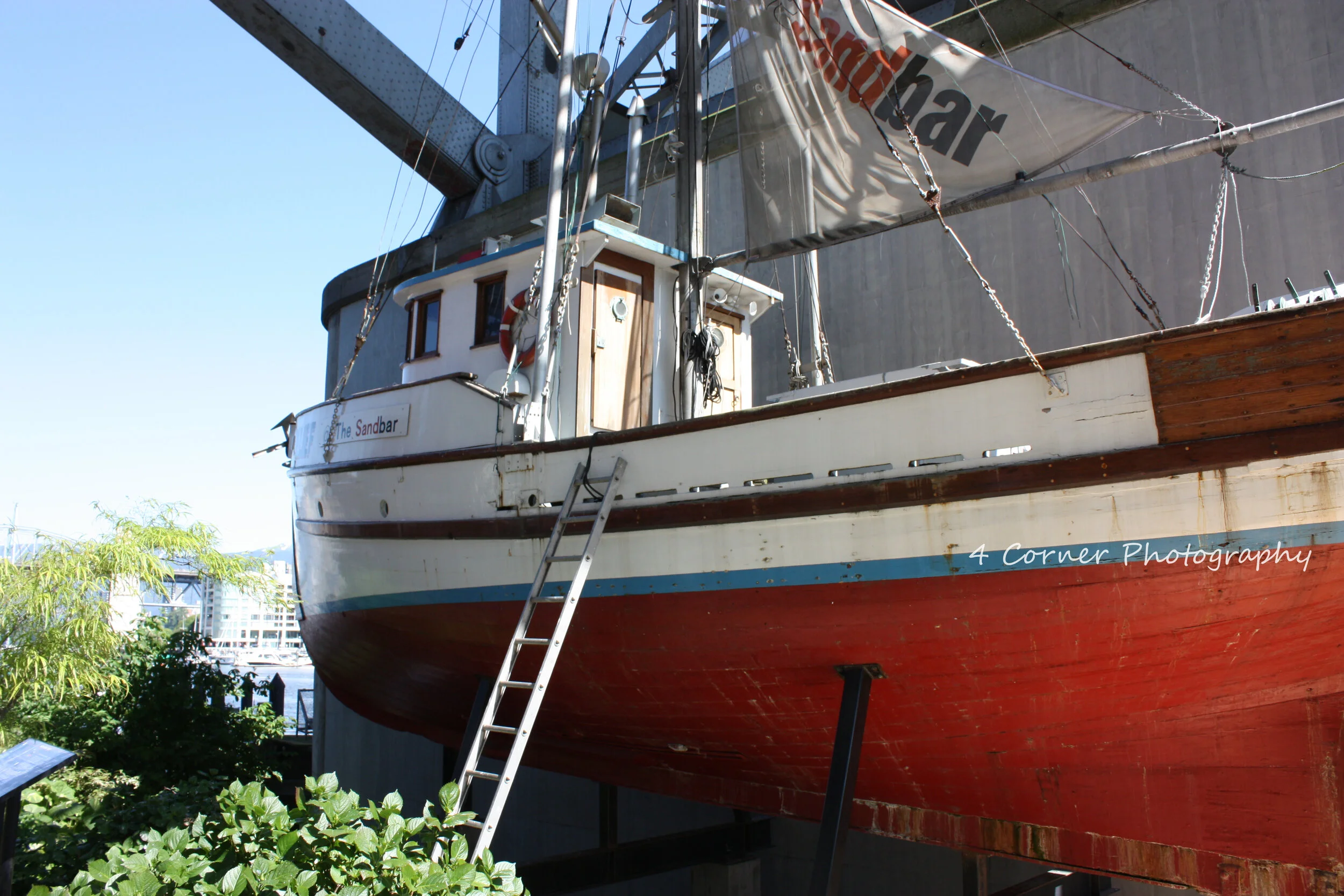 A wooden sailboat named The Sandbar is displayed on land, supported by stands, with a ladder leaning against its hull. The boat has a white and red hull, with a small cabin and a sail partially visible.