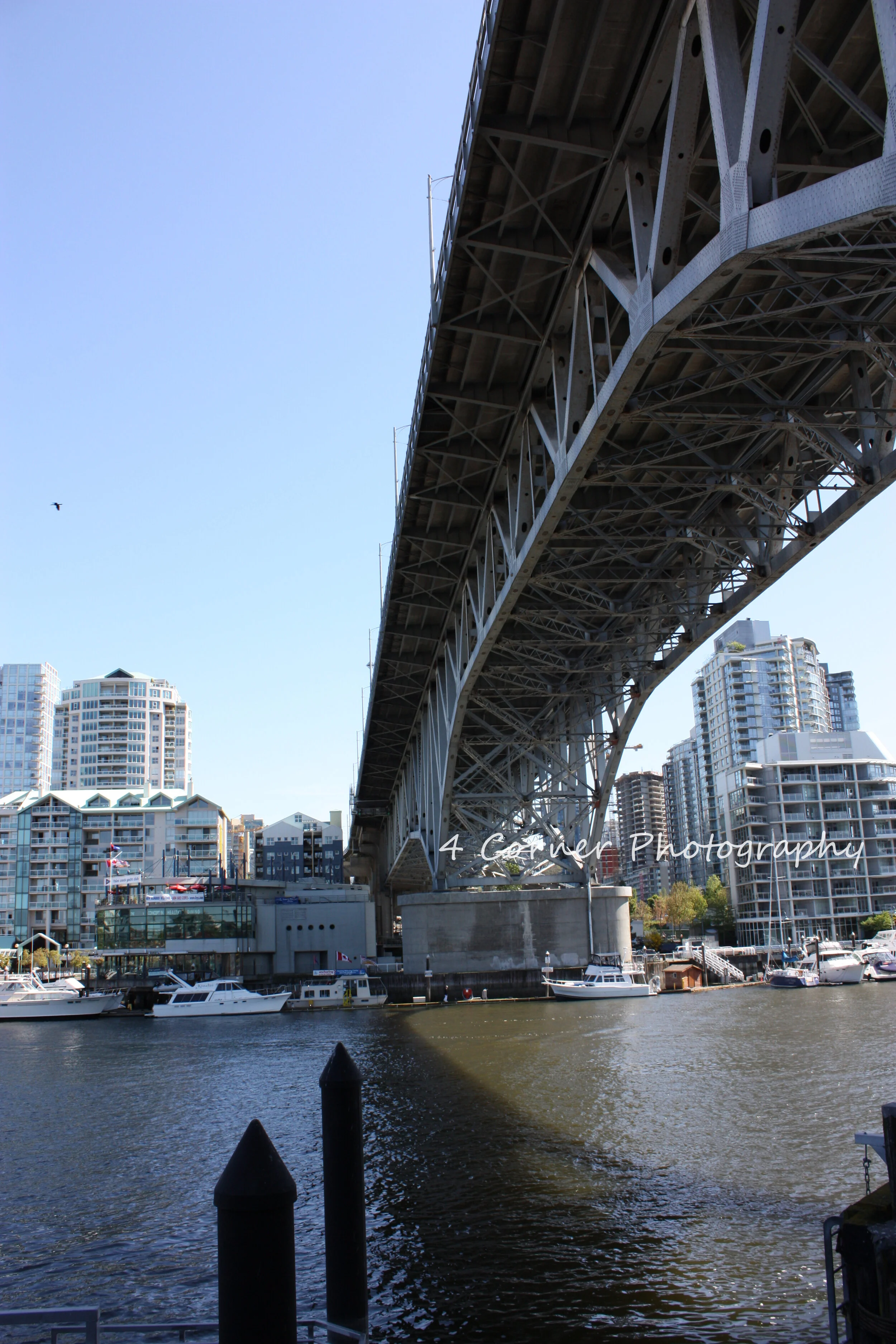 Underneath a large steel bridge over a marina with boats and modern high-rise buildings in the background.
