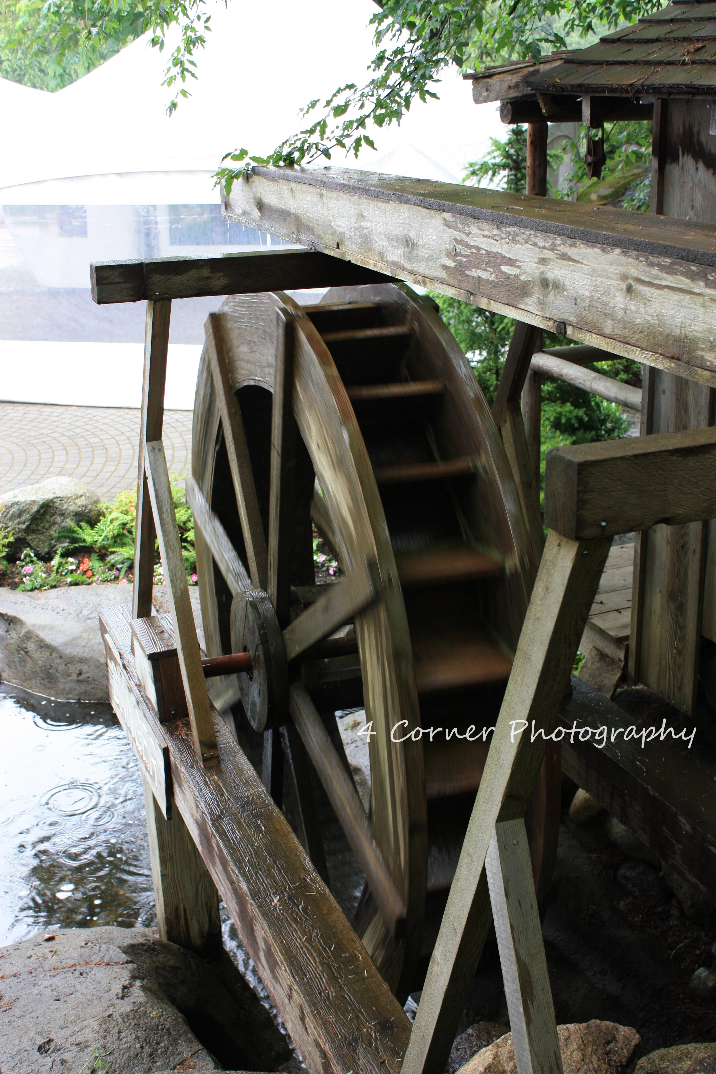 A wooden water wheel attached to a small water canal, with greenery and part of a building in the background.