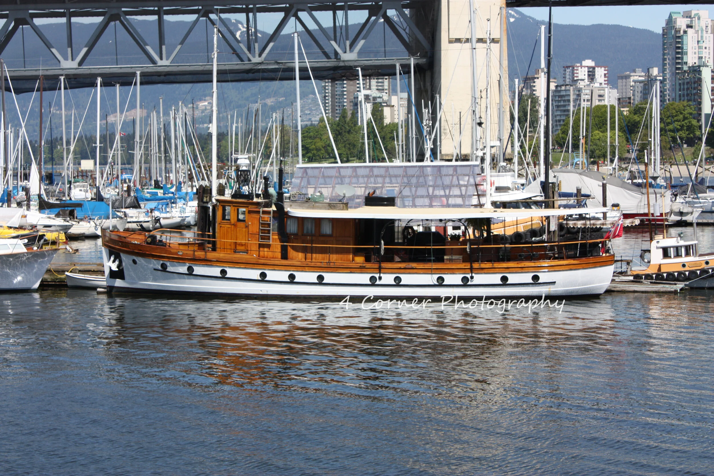 A large wooden and white motorboat docked at a marina, with numerous sailboats and yachts in the background, a bridge overhead, and a cityscape with high-rise buildings and forested hills in the distance.