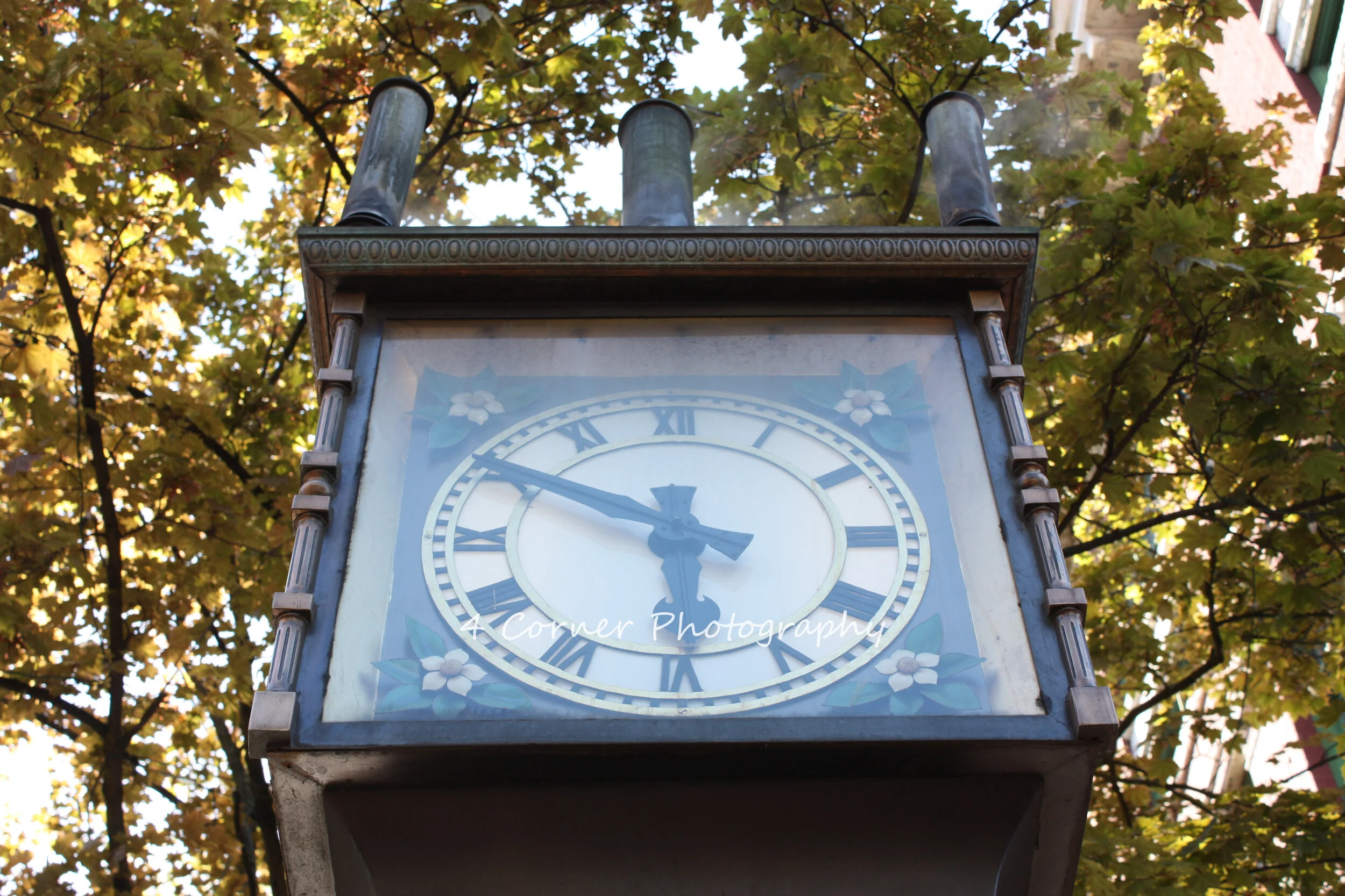 An outdoor vintage clock with Roman numerals, showing the time at approximately 4:47, mounted on a metal structure against a background of green and yellow autumn leaves on trees.