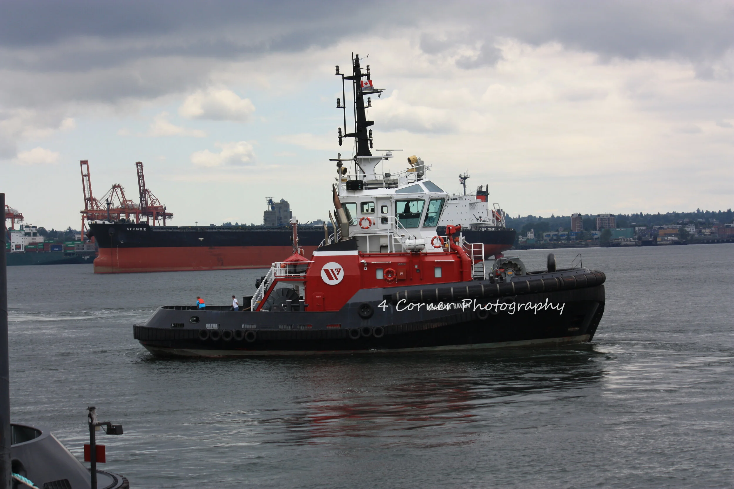 A red and black tugboat on the water with a larger cargo ship in the background under a mostly cloudy sky.