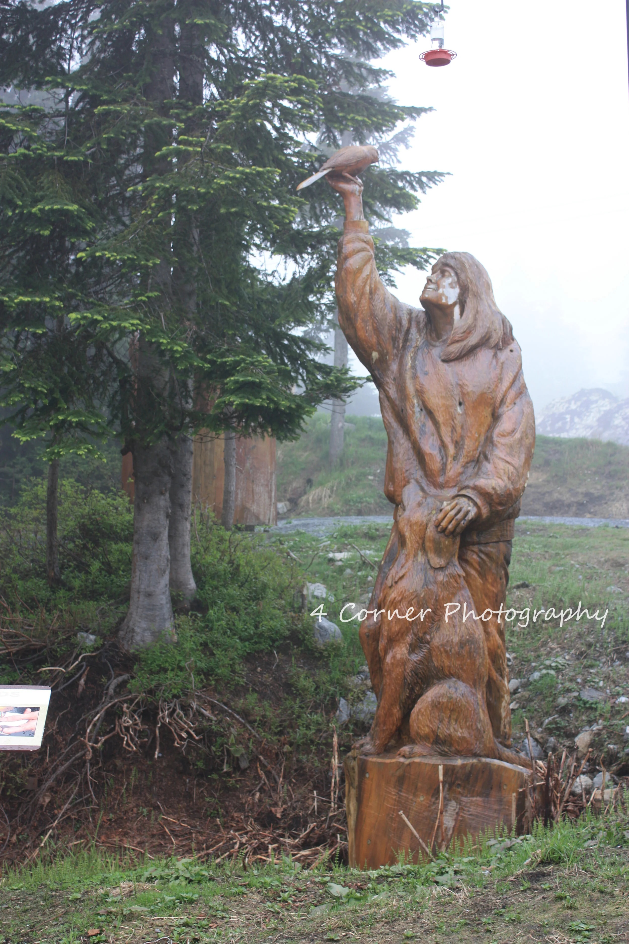 Wooden sculpture of a woman holding a bird, situated outdoors among trees and greenery.