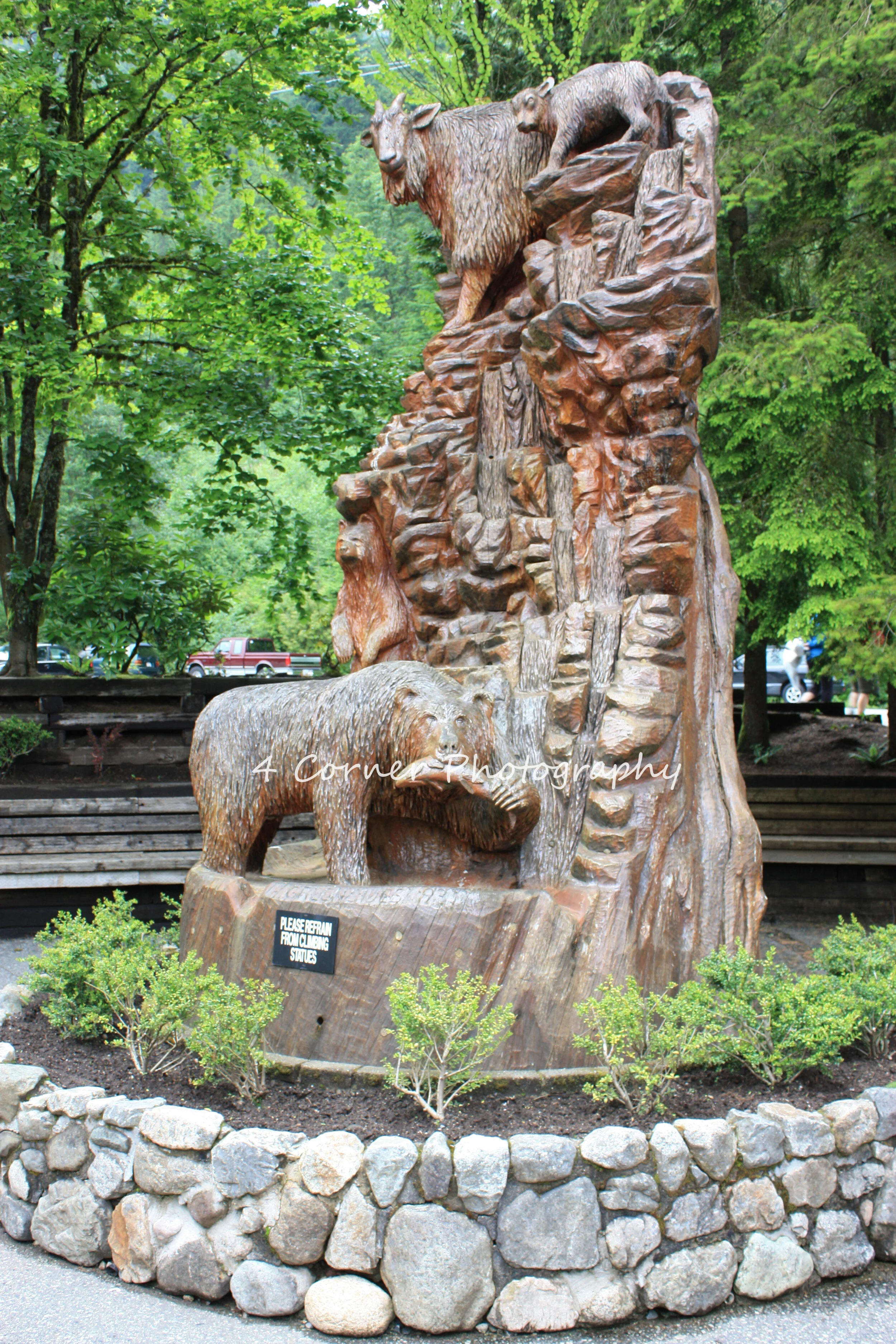 Wooden sculpture of a bear, a mountain lion, a moose, and a bison in a wooded park setting with trees and greenery.