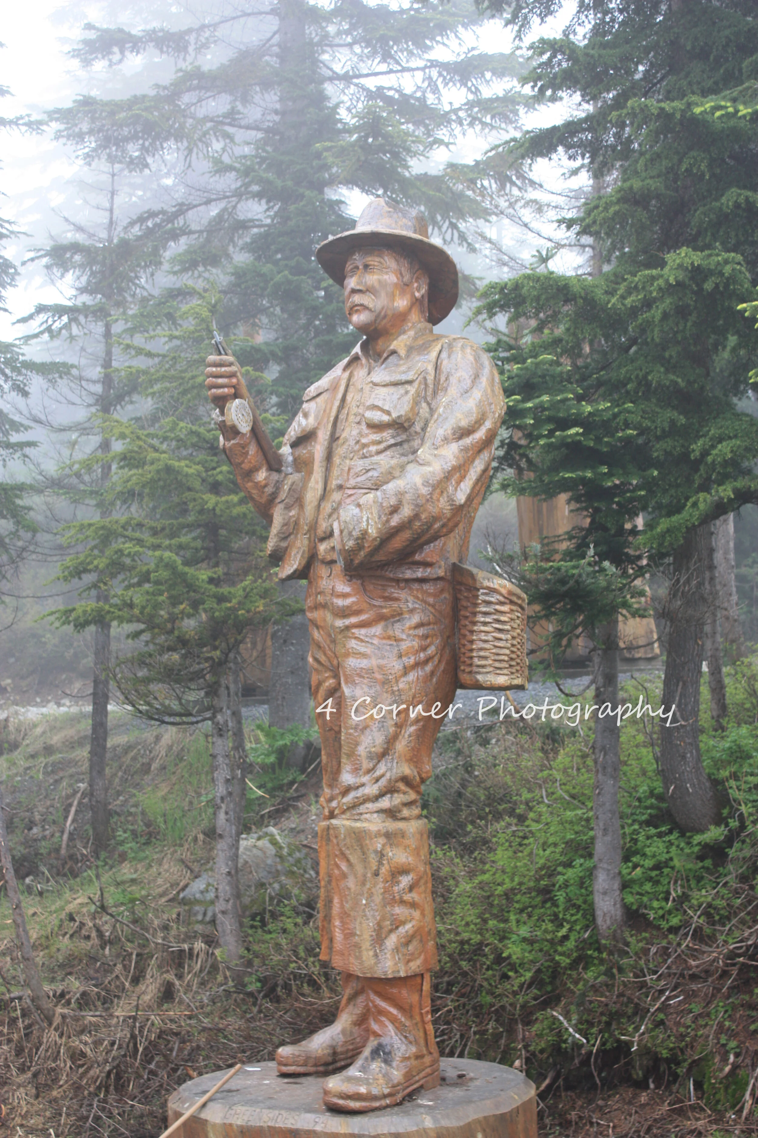 Wooden statue of a man wearing a hat, standing outdoors among trees, holding a cell phone, with a basket on his hip.