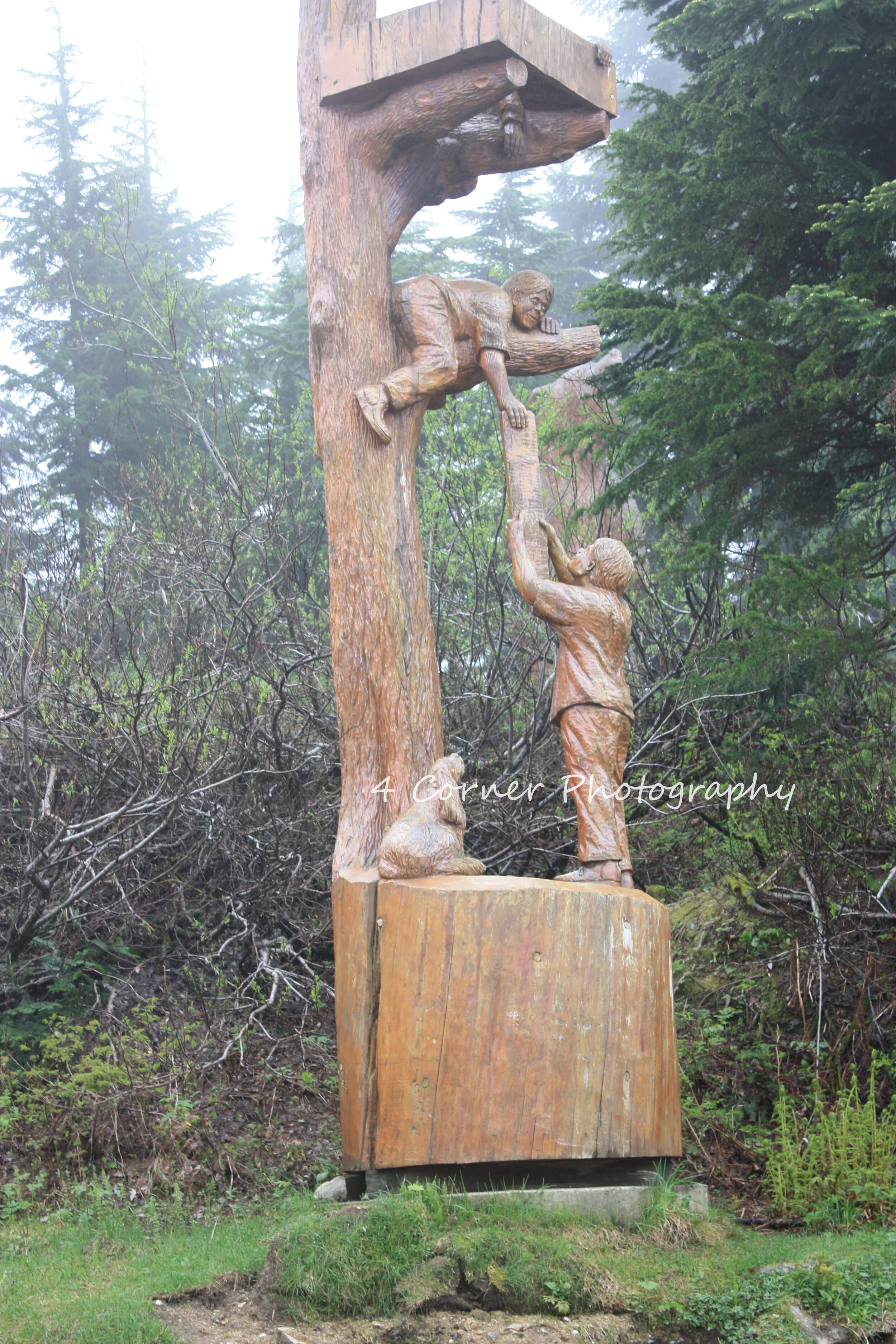 Wooden sculpture of a boy reaching down to a girl sitting on a log, with a dog at the base, set in an outdoor forested area with trees and mist.