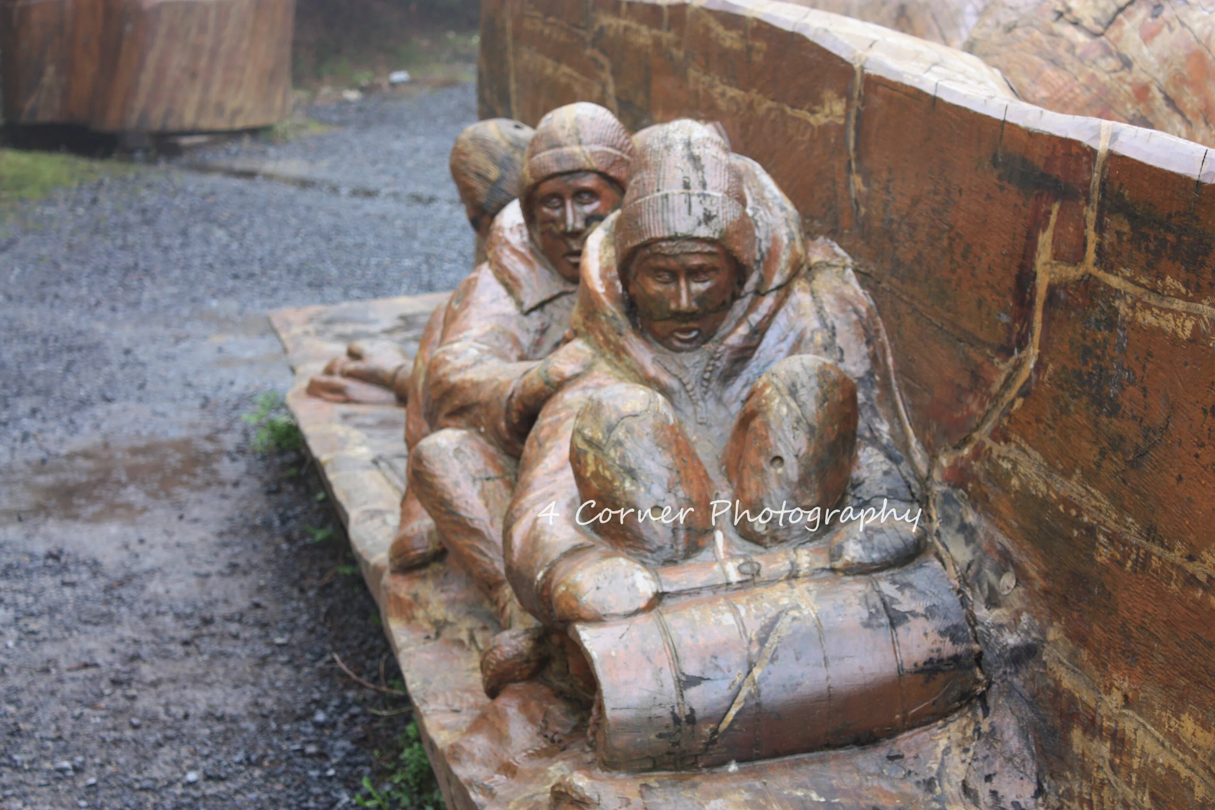 Wooden sculpture of three people lying on the ground, close to a brick wall, with wet ground nearby, by 4 Corner Photography.