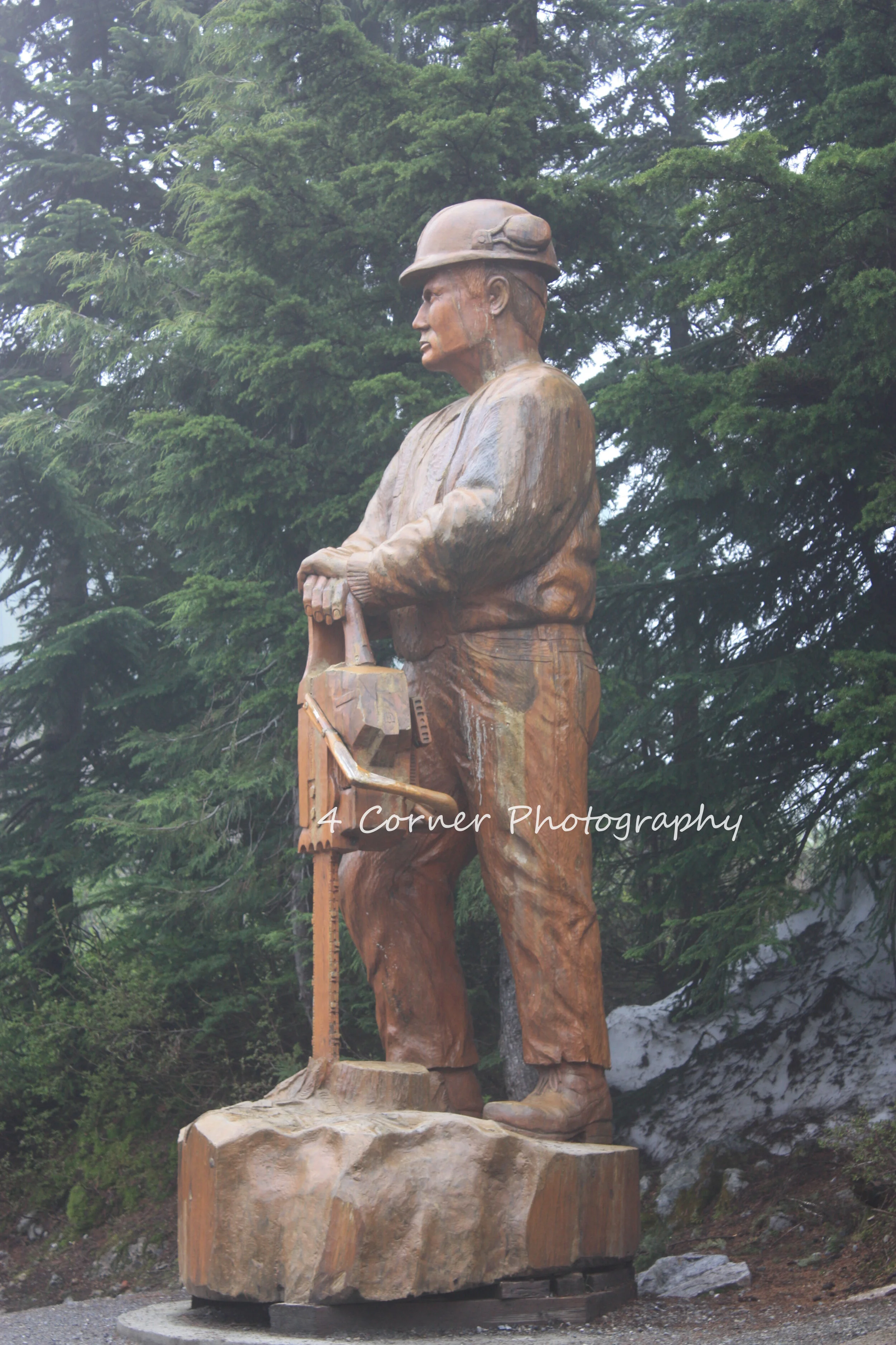 Wooden statue of a man wearing a hard hat and work clothes, holding a chainsaw, set against a background of trees.