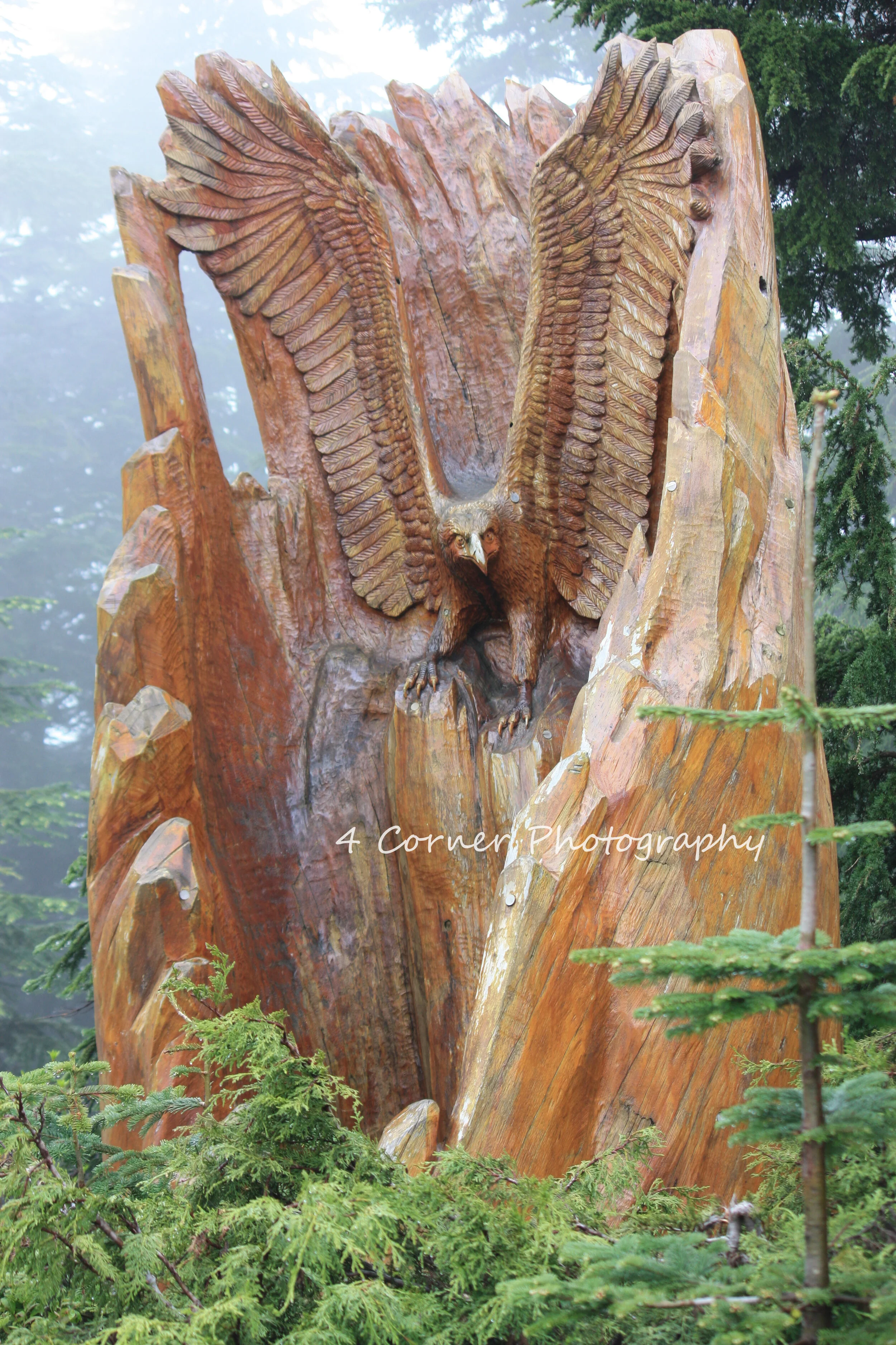Wood carving of an eagle with wings spread, carved into a large tree trunk. The carving is set outdoors among green trees.