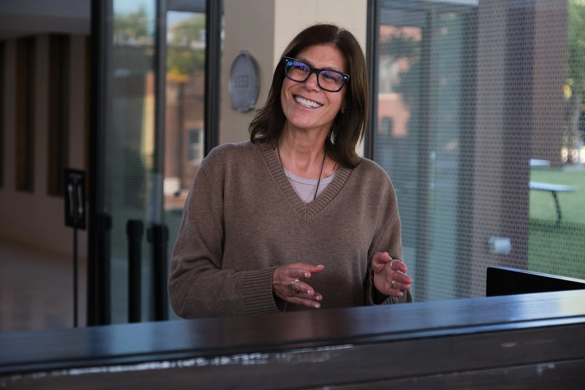 A woman with shoulder-length brown hair and glasses smiling and gesturing with her hands.