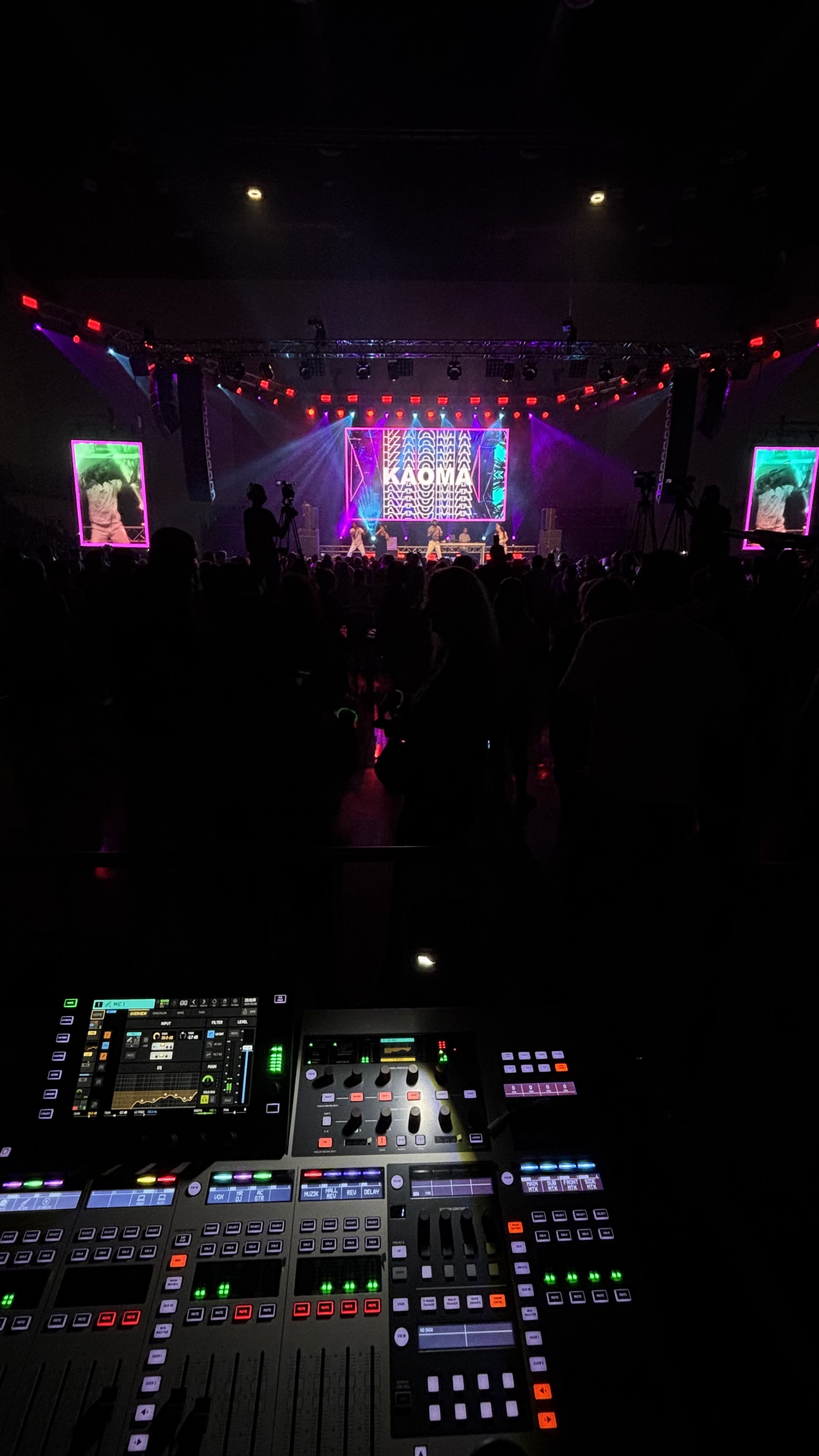 View from behind sound and lighting control panel at a concert or event with silhouettes of audience, stage lights, performers, and digital screens displaying colorful visuals.