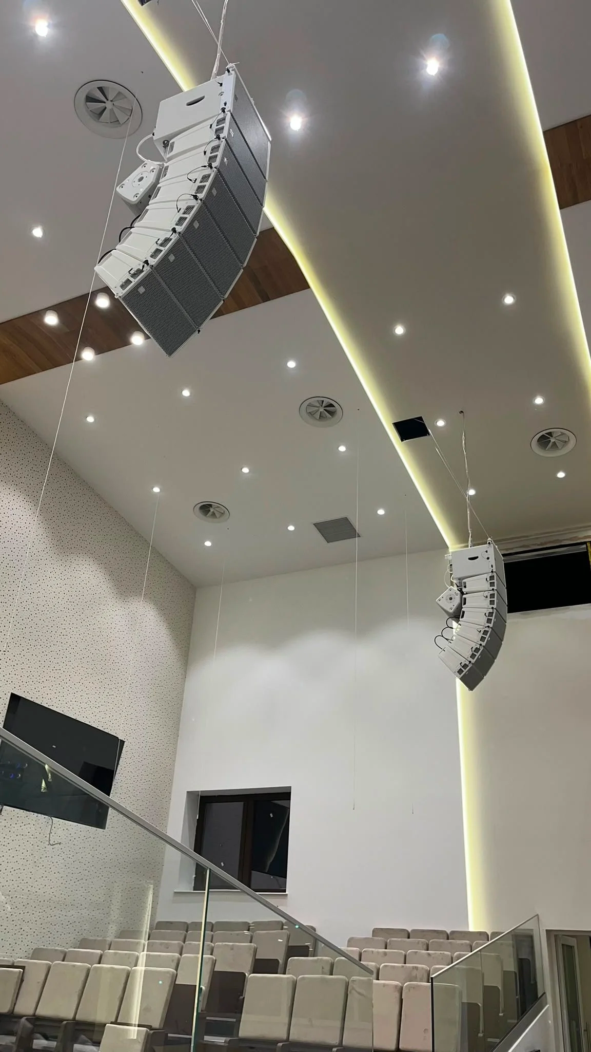 Interior of a modern auditorium or church with beige seating, white walls, round ceiling lights, and large speaker systems hanging from the ceiling.