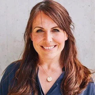 Smiling woman with long brown hair, wearing a blue shirt and a round pendant necklace, against a gray background.