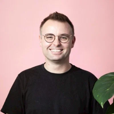 Smiling person with short hair and glasses in front of a pink background, wearing a black shirt and standing next to green plant leaves.