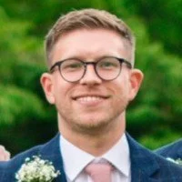 A smiling man wearing glasses, a suit jacket, and a tie, with a blurred green background.