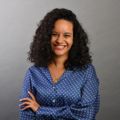 A smiling person with curly hair, wearing a blue patterned blouse, standing against a plain background.