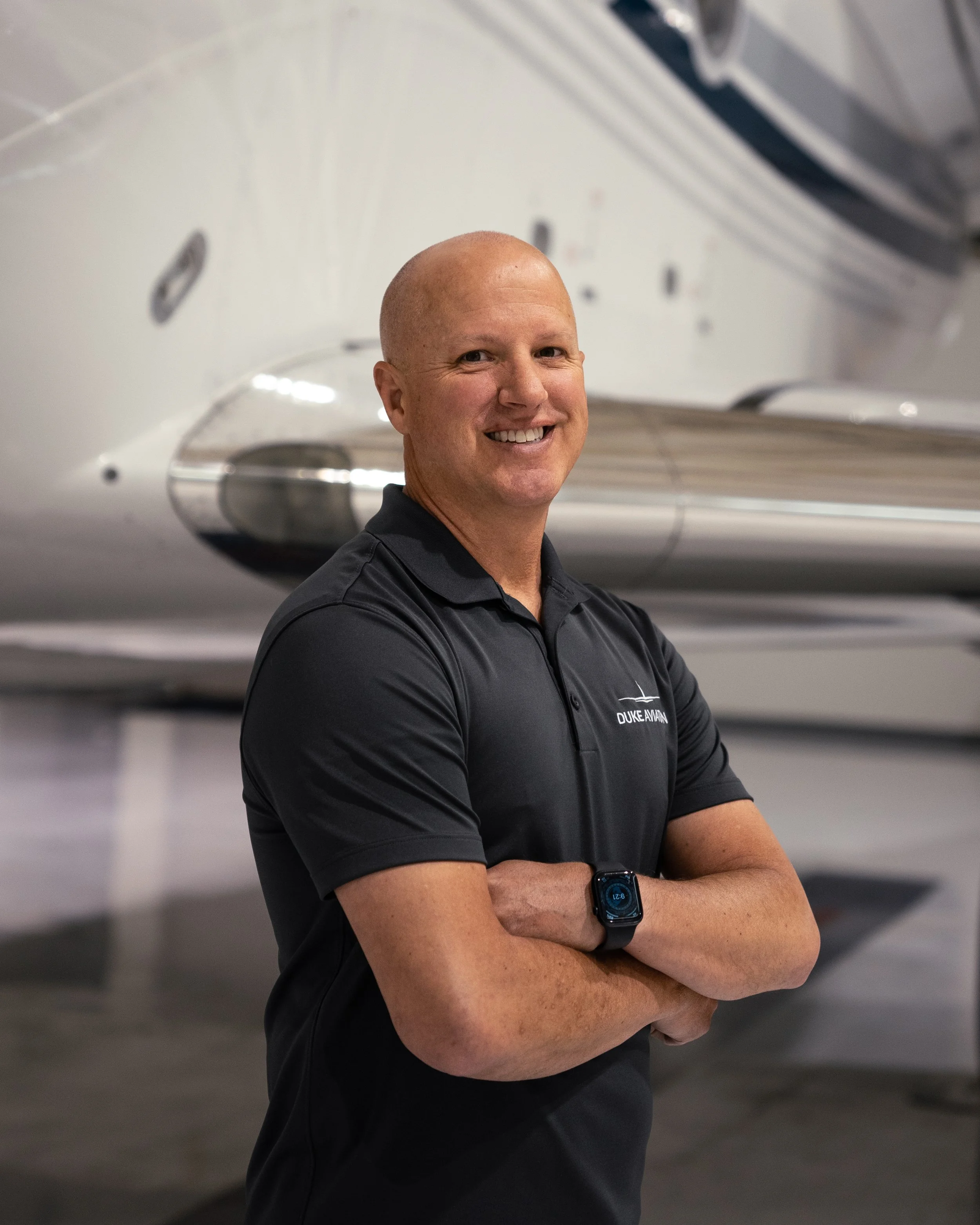 Man in a dark polo shirt standing with arms crossed in front of an airplane in a hangar.