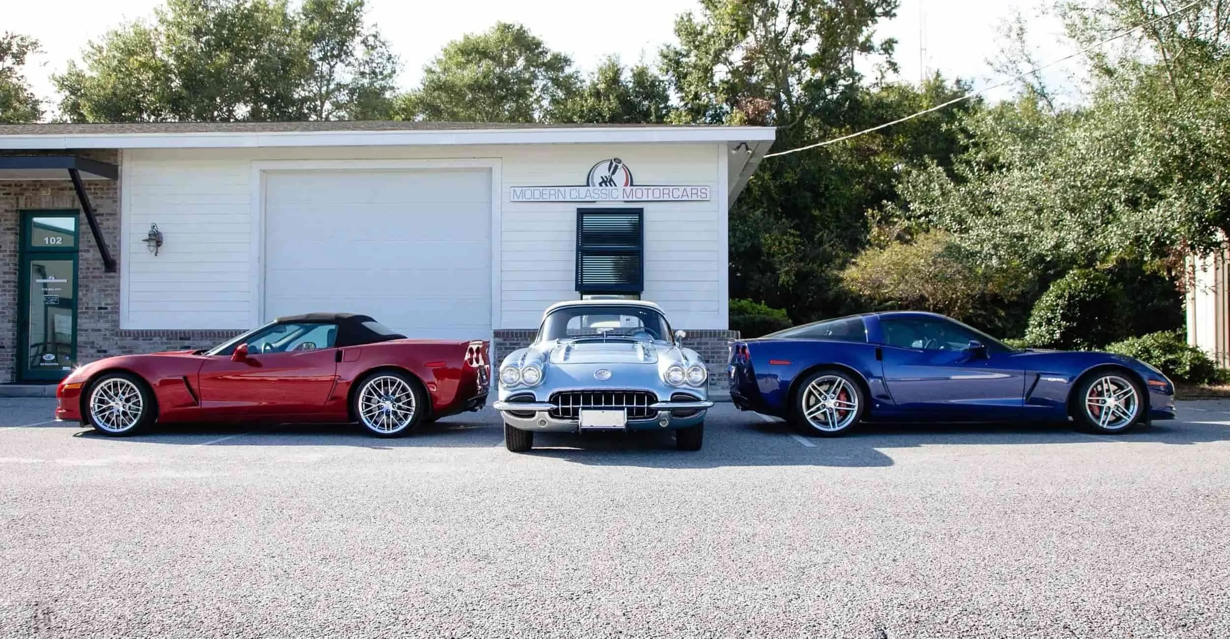 A red Corvette, a classic silver Corvette, and a blue Corvette parked side by side in front of a white building.