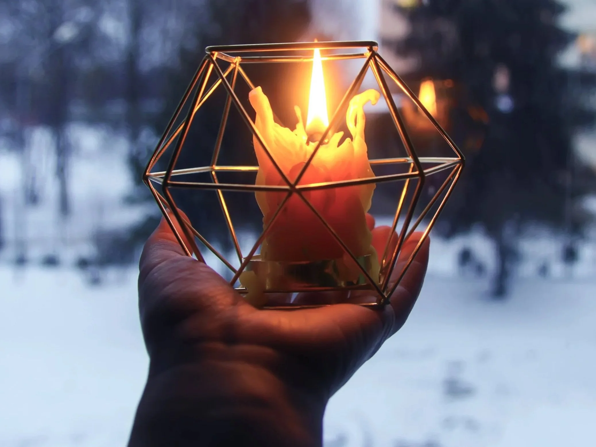 An outstretched hand holds a glowing beeswax candle in a geometric holder at dusk, surrounded by snow and trees in an urban forest, symbolizing the Archer’s devotion and the light of the New Moon in Sagittarius and the Winter Solstice.