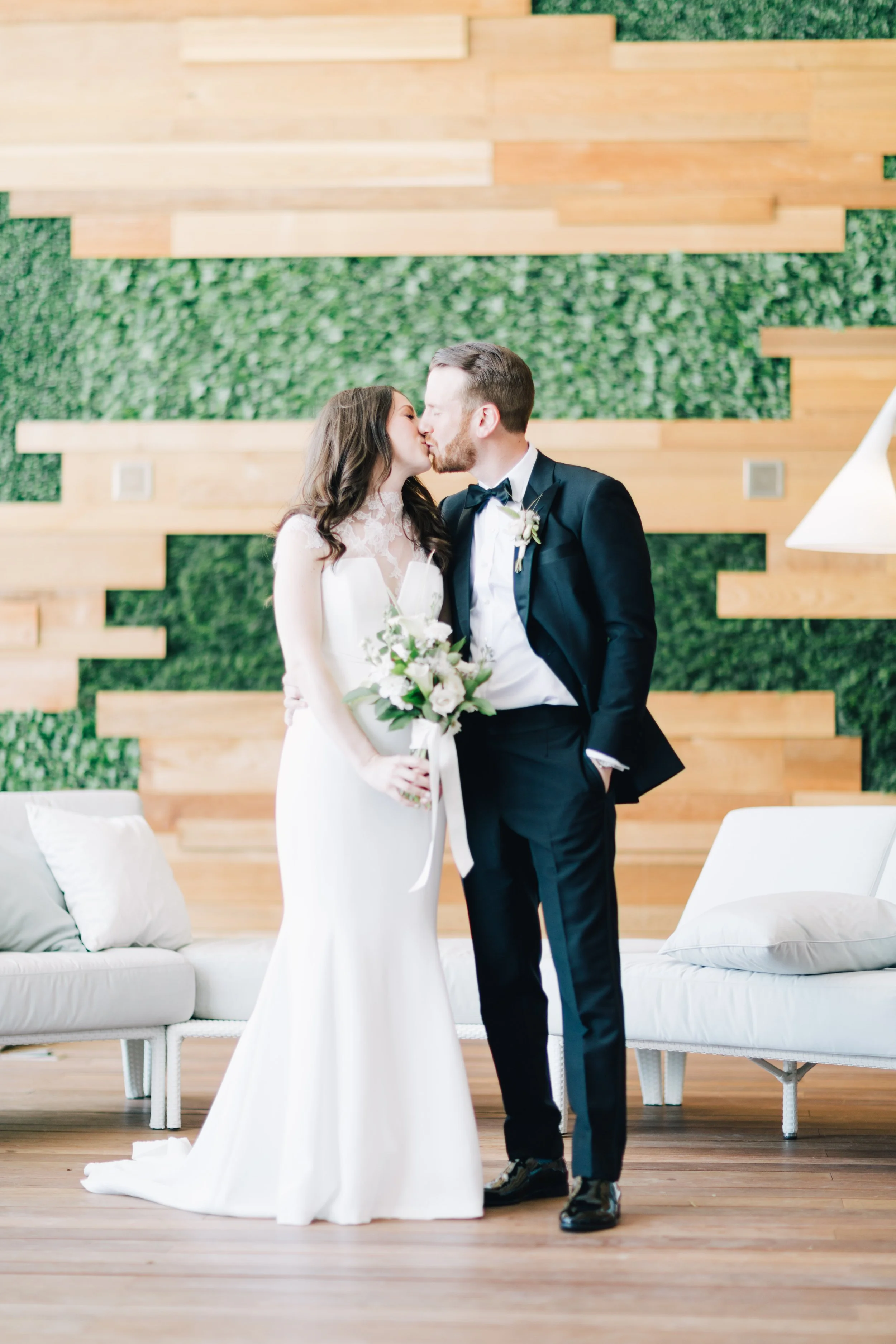 A bride and groom sharing a kiss at their wedding, standing in front of a modern wood and green wall, with a white sofa nearby.