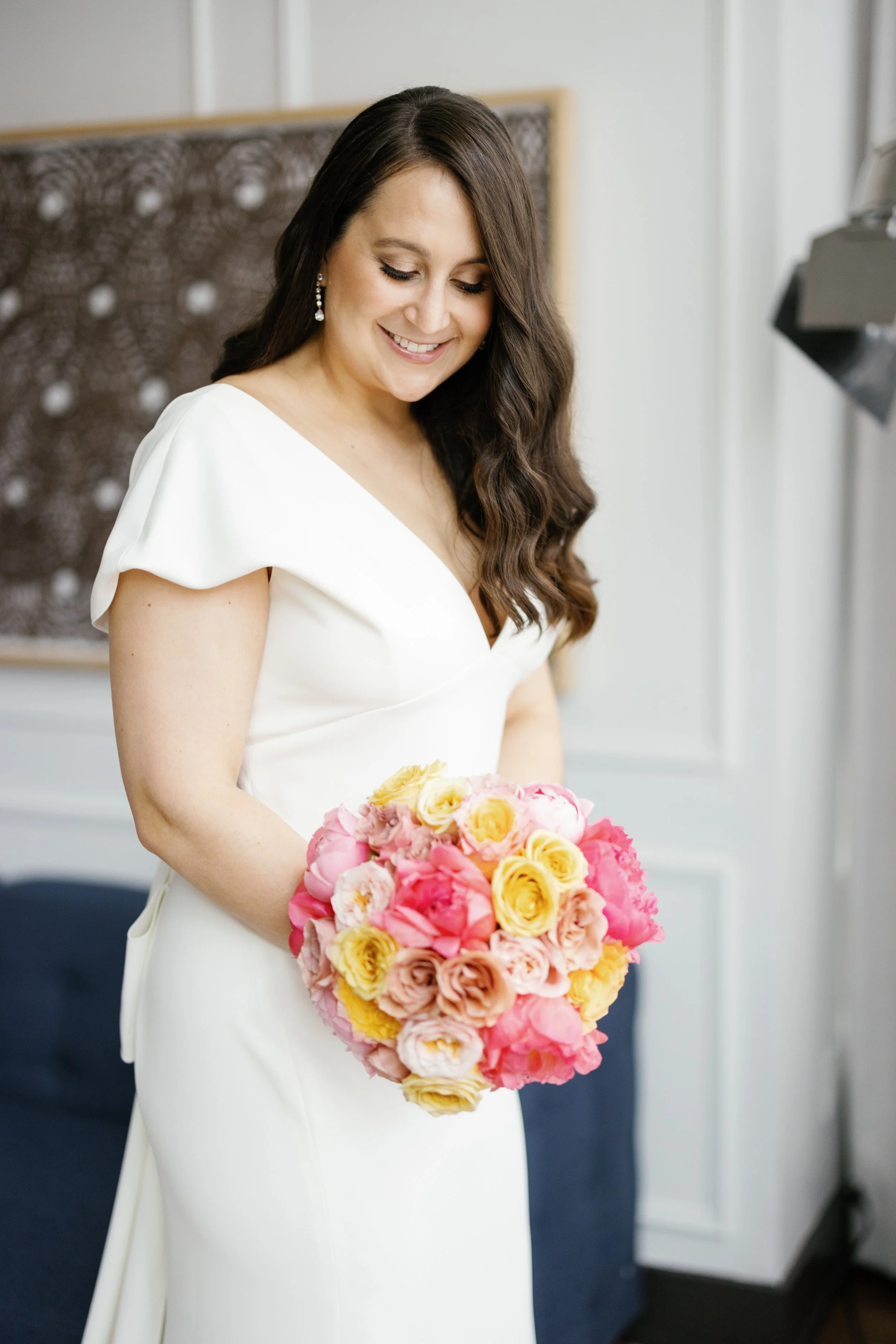 Woman in white dress holding a bouquet of pink and yellow roses smiling with long brown hair and earrings in a room with art on the wall.
