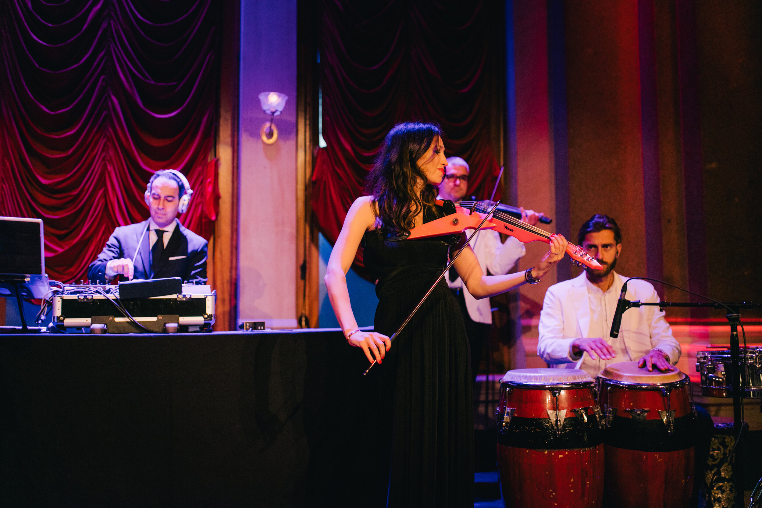 A woman playing a violin on stage while a band accompanies her, with red curtains in the background.
