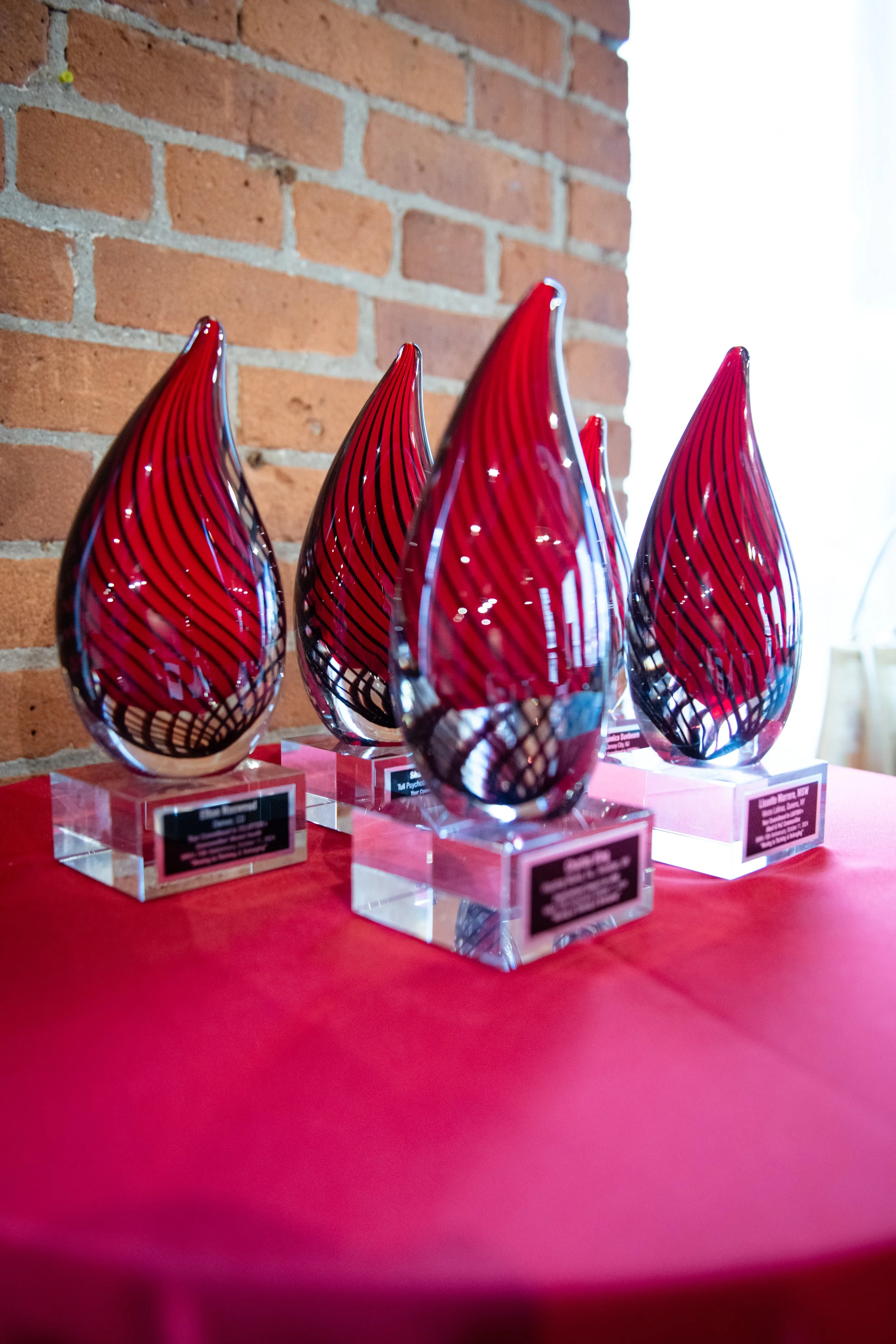 Close-up of four decorative glass art pieces with red and black swirled patterns, displayed on clear acrylic stands on a red surface against a brick wall background.
