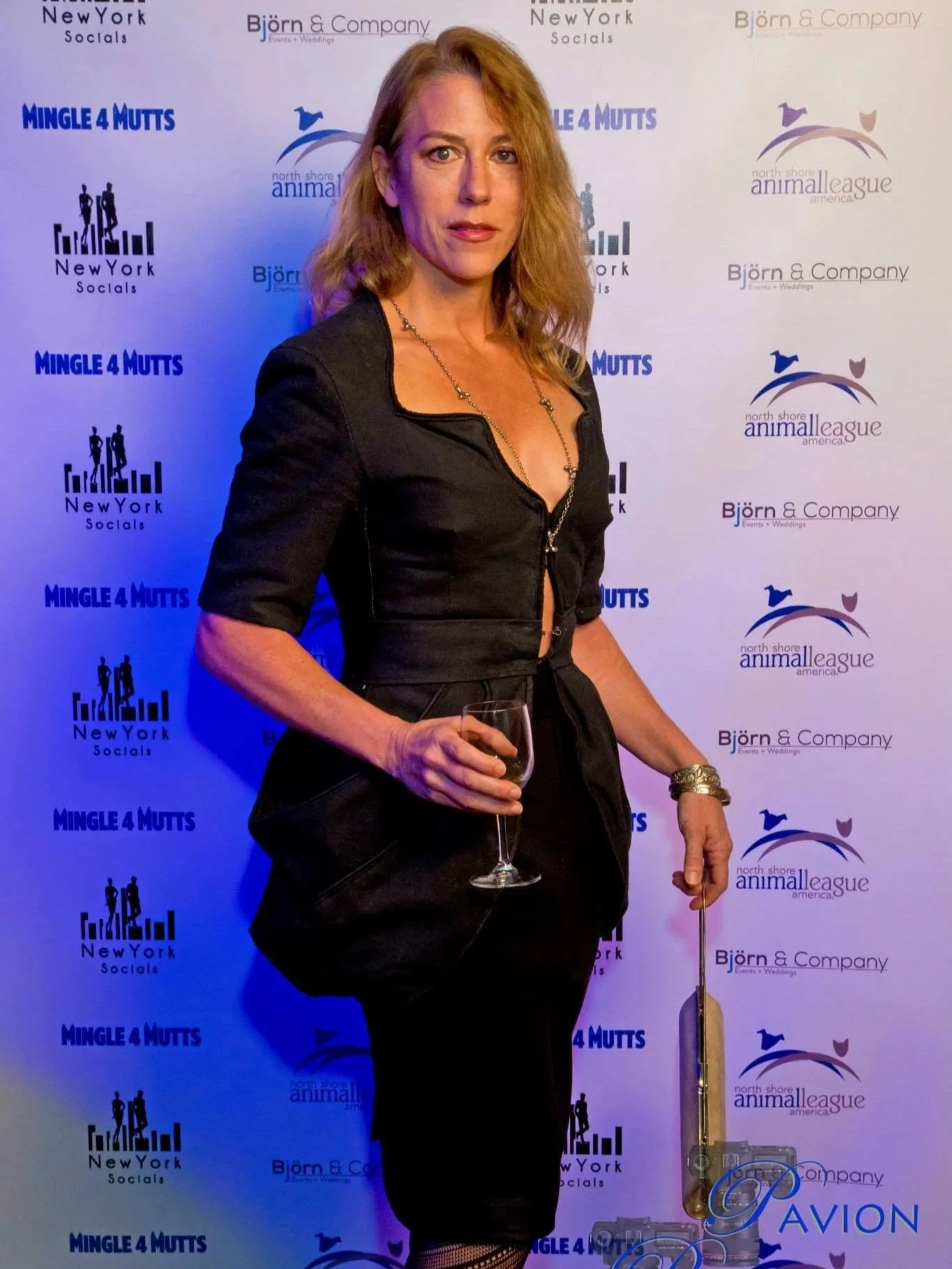 A woman holding a glass of white wine at an event with a backdrop featuring logos of organizations including Mingle 4 Mutts, North Shore Animal League America, and Björn & Company.